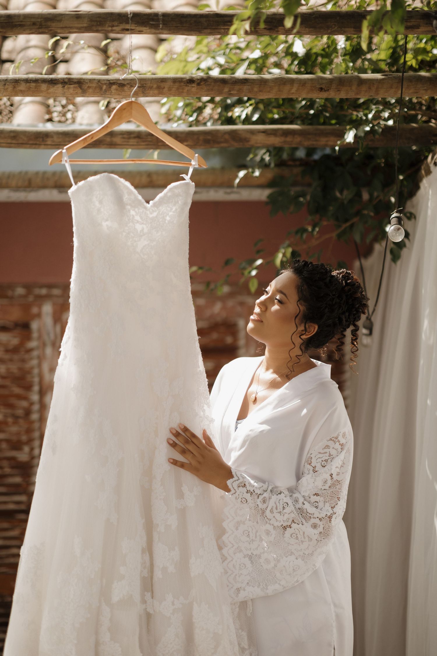 A beautiful white wedding dress hangs elegantly against a rustic brick wall with green foliage in the background.