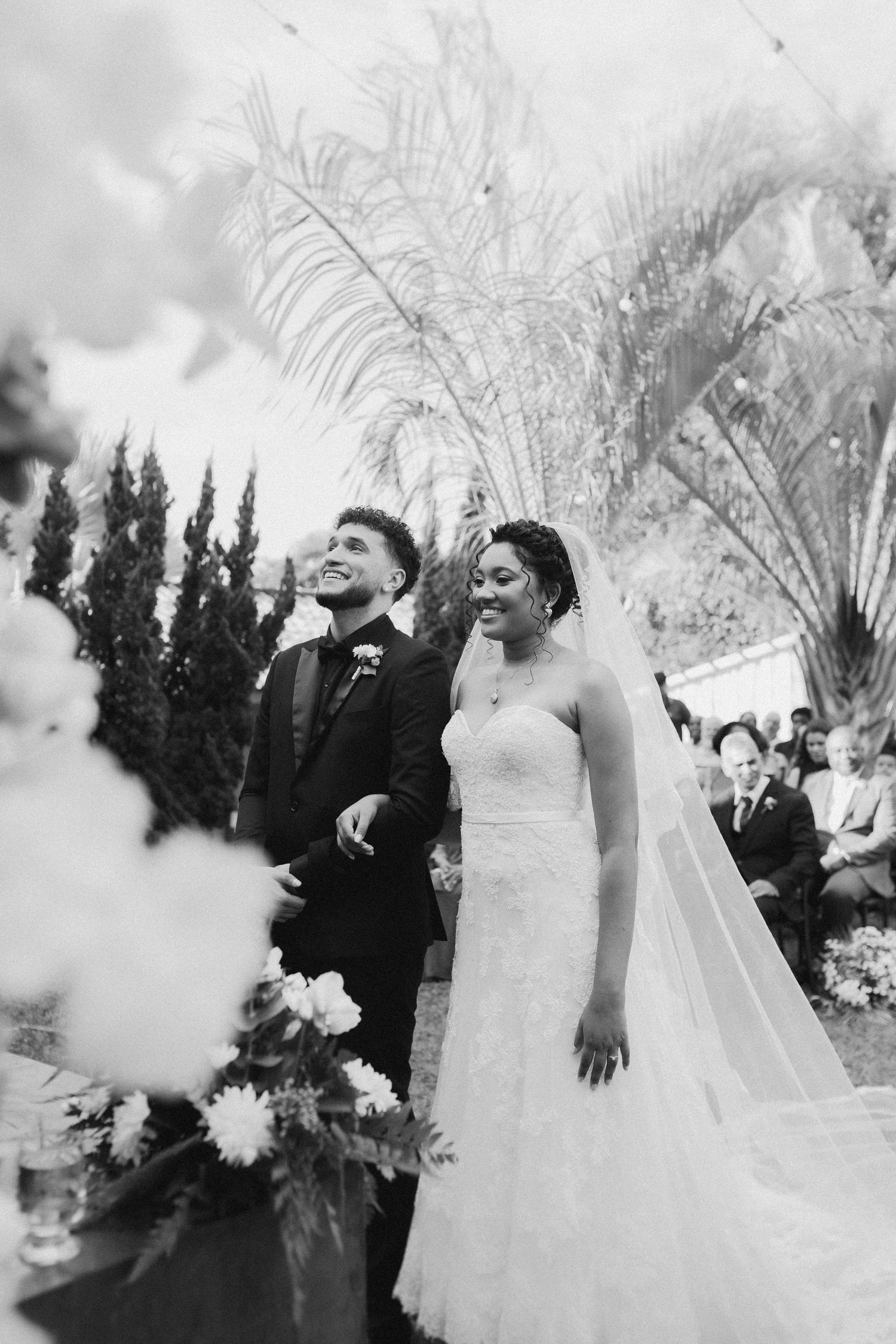 Artistic black and white wedding photo with palm trees and evergreens in the background.