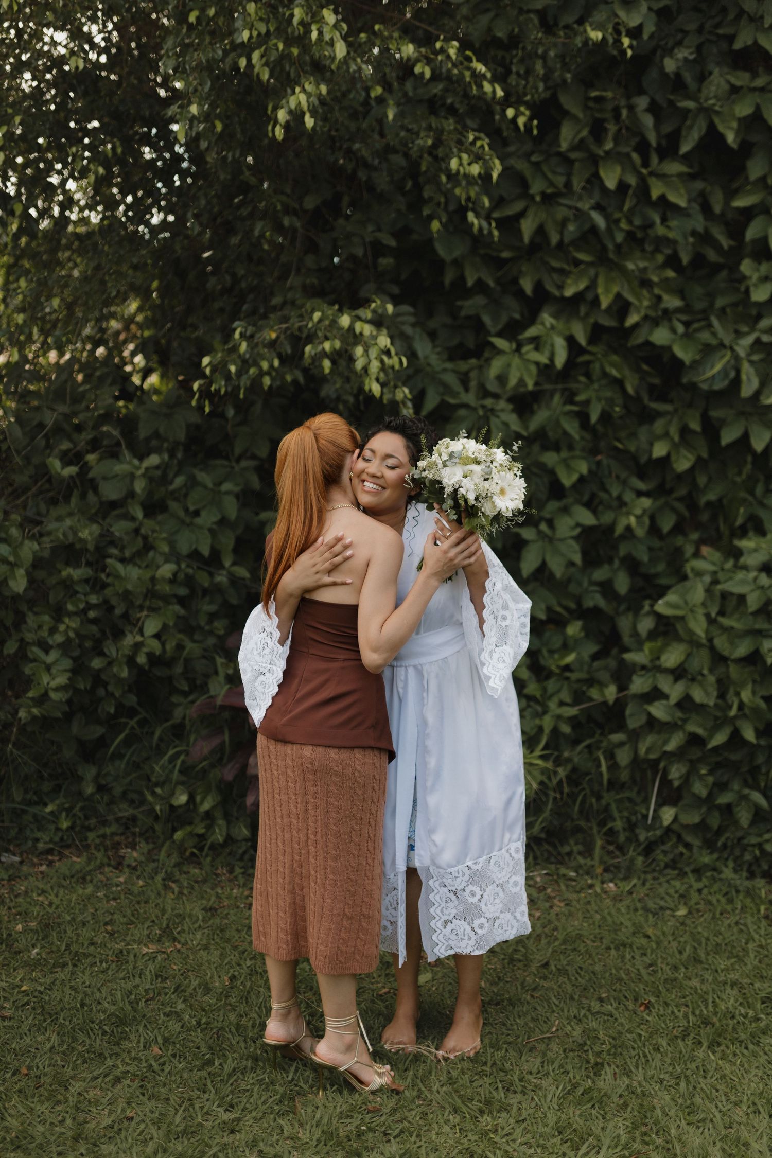 Two people embrace warmly outdoors, one in a brown dress and one in white lace.
