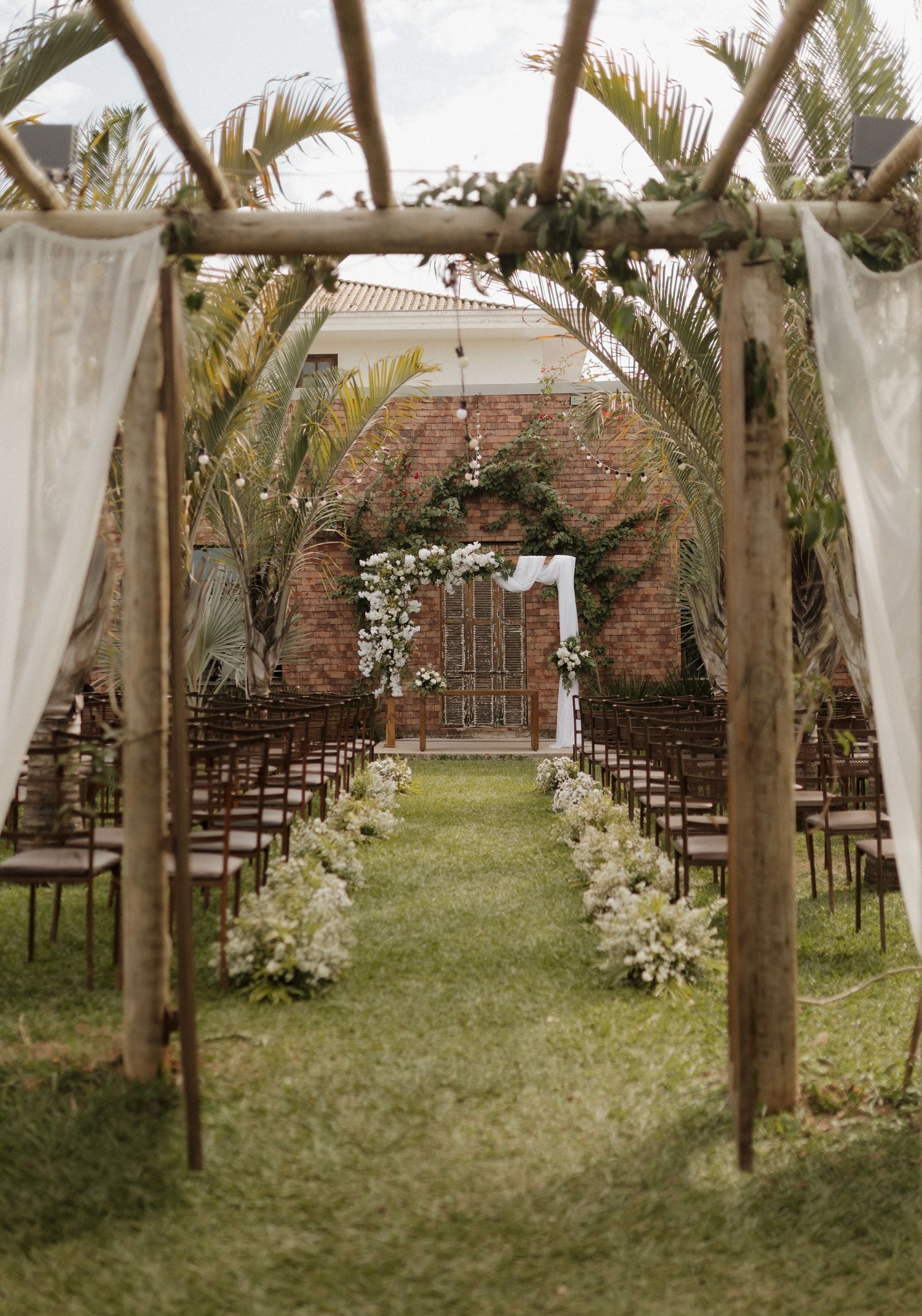 Outdoor wedding ceremony setup with white drapes and palm trees lining a grass aisle.