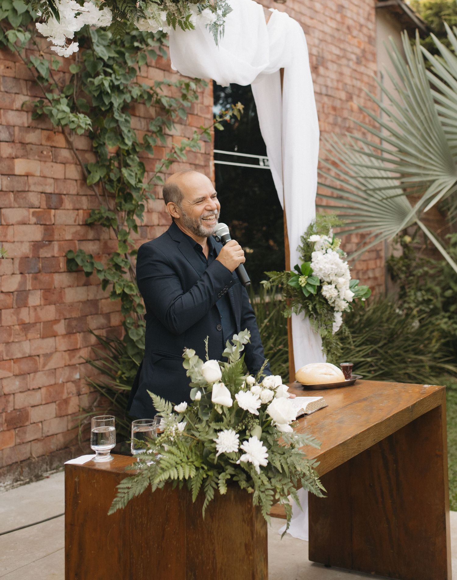 A speaker stands at a wooden podium decorated with white flowers against a brick wall.