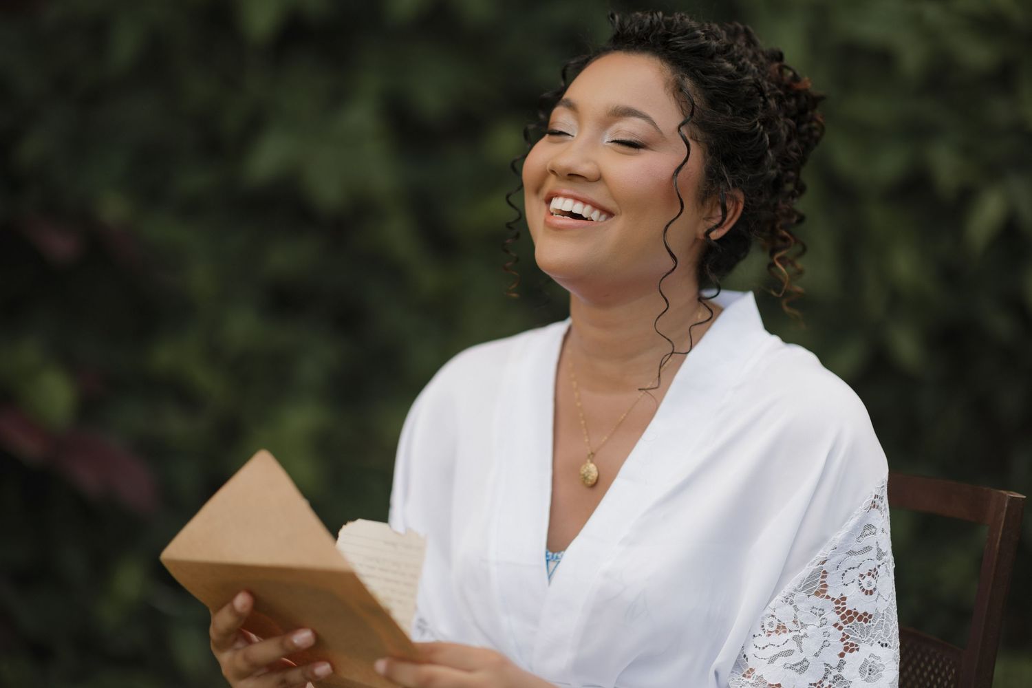 A person in a white lace robe laughs while sitting in a garden setting.