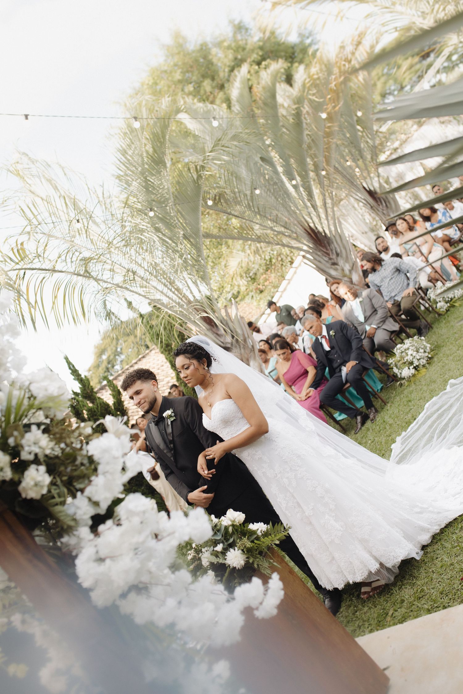 Wedding ceremony scene with guests seated outdoors under palm trees and white floral decorations.