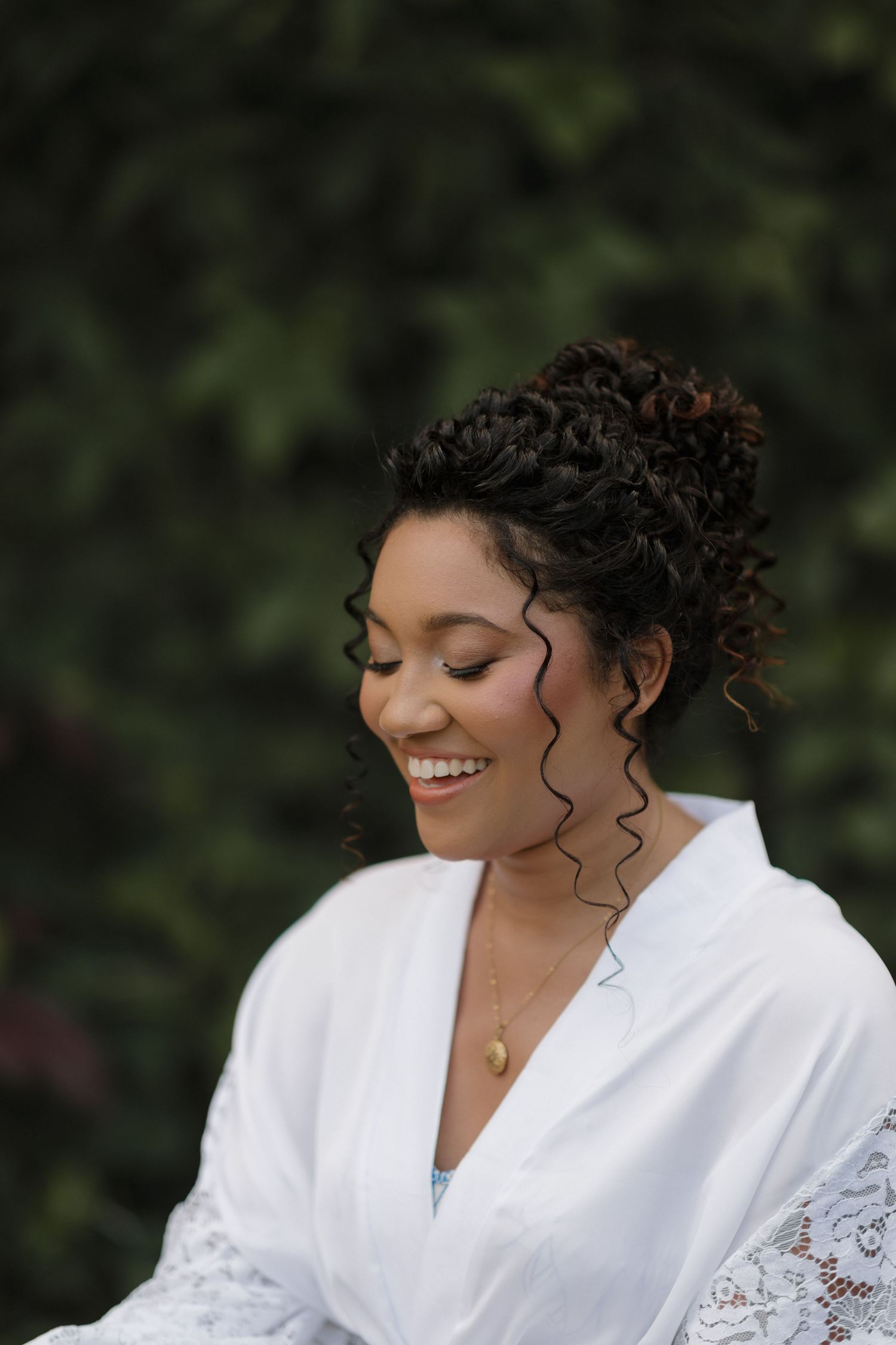 A person in a white lace robe smiles joyfully while sitting outdoors against a natural green backdrop.