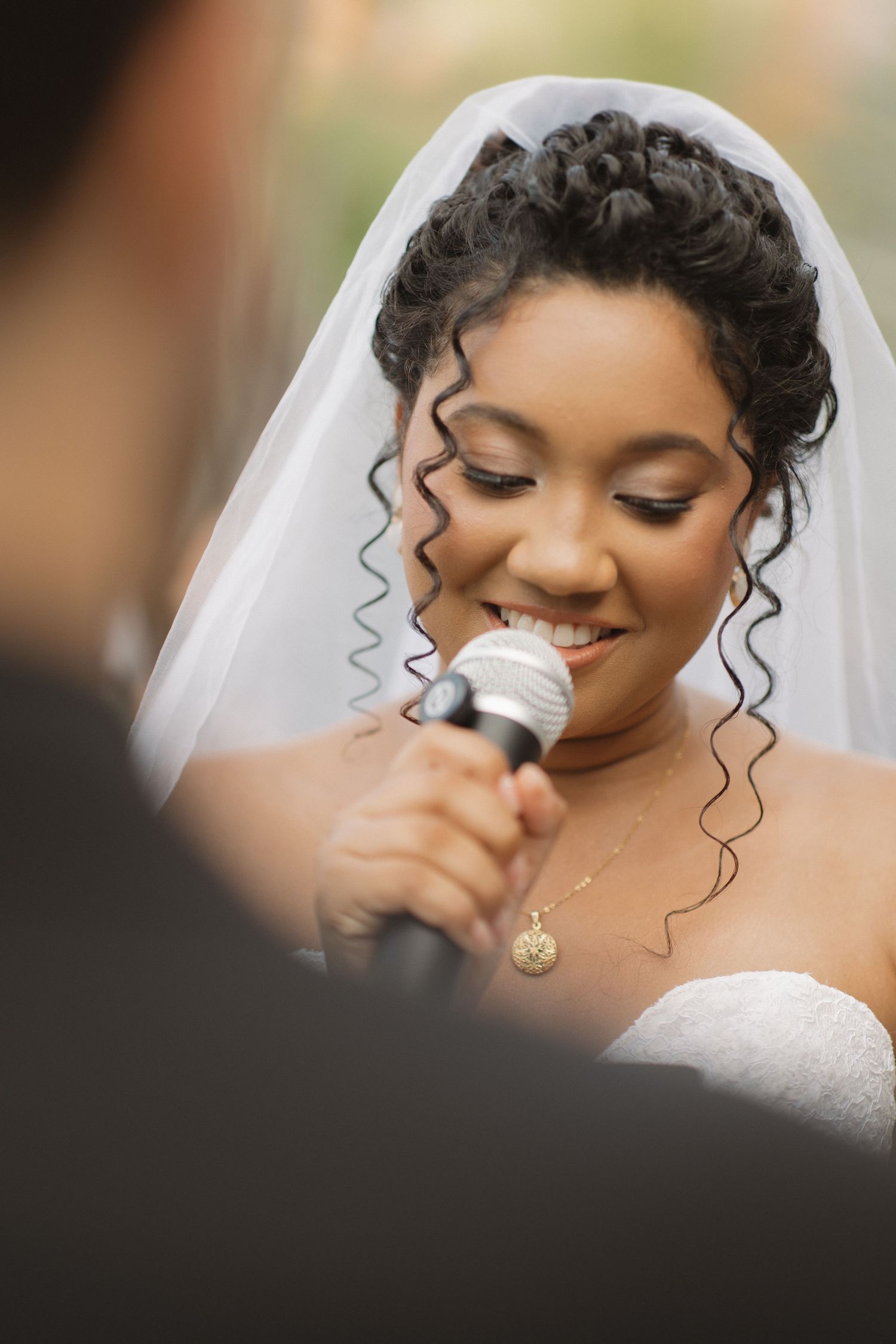A bride wearing a delicate veil and necklace smiles during her wedding ceremony.