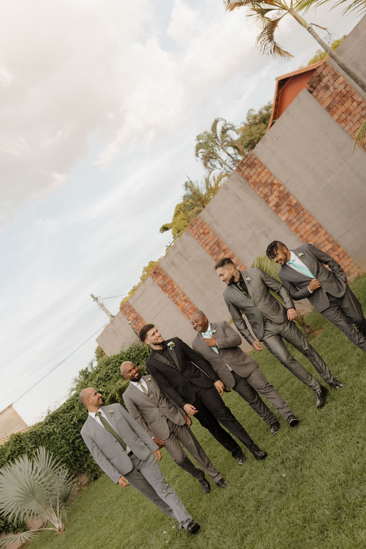 Groomsmen in gray suits pose casually on lawn near brick wall.