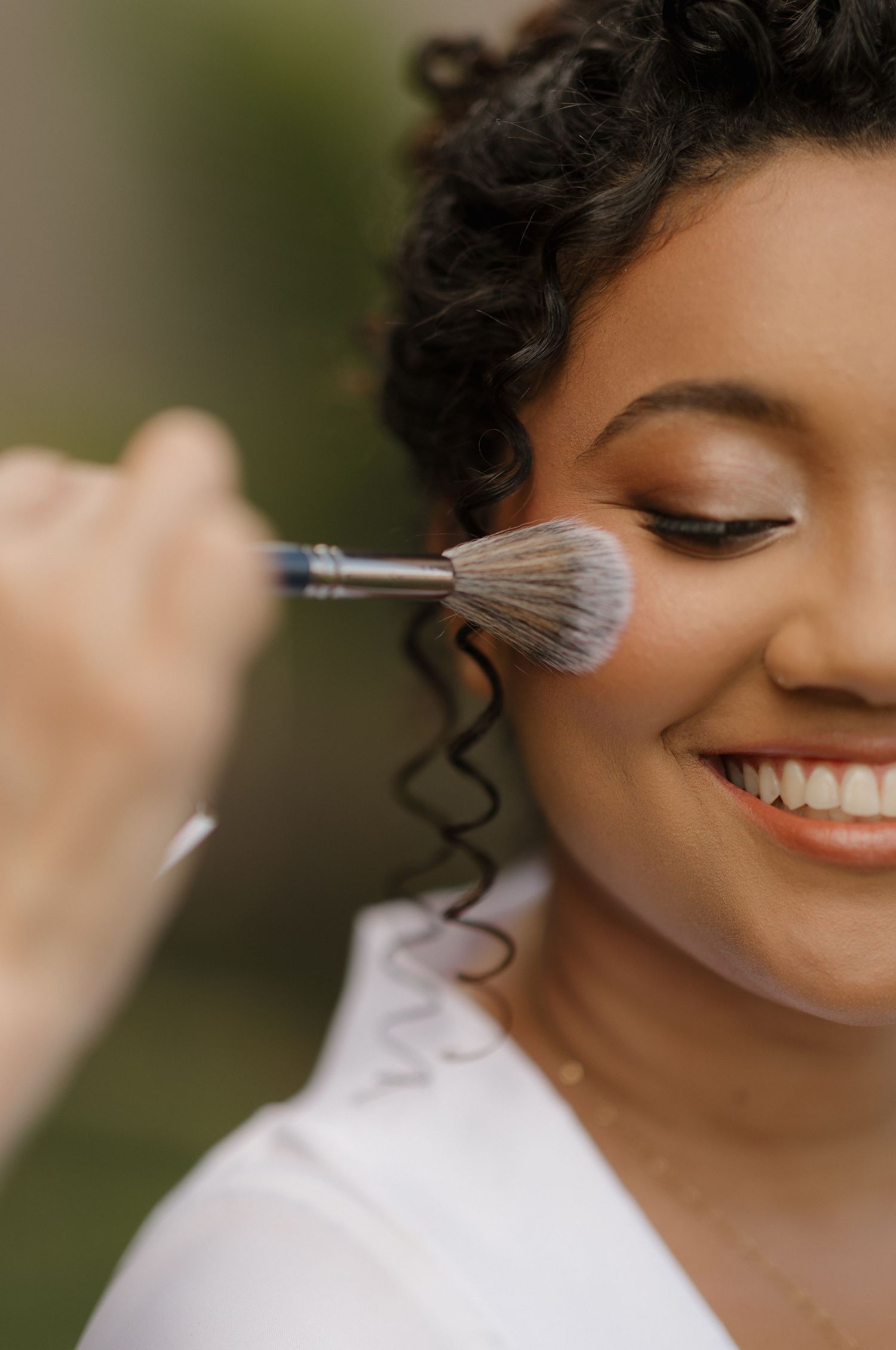 A makeup brush applies cosmetics during bridal preparation against a natural outdoor background.
