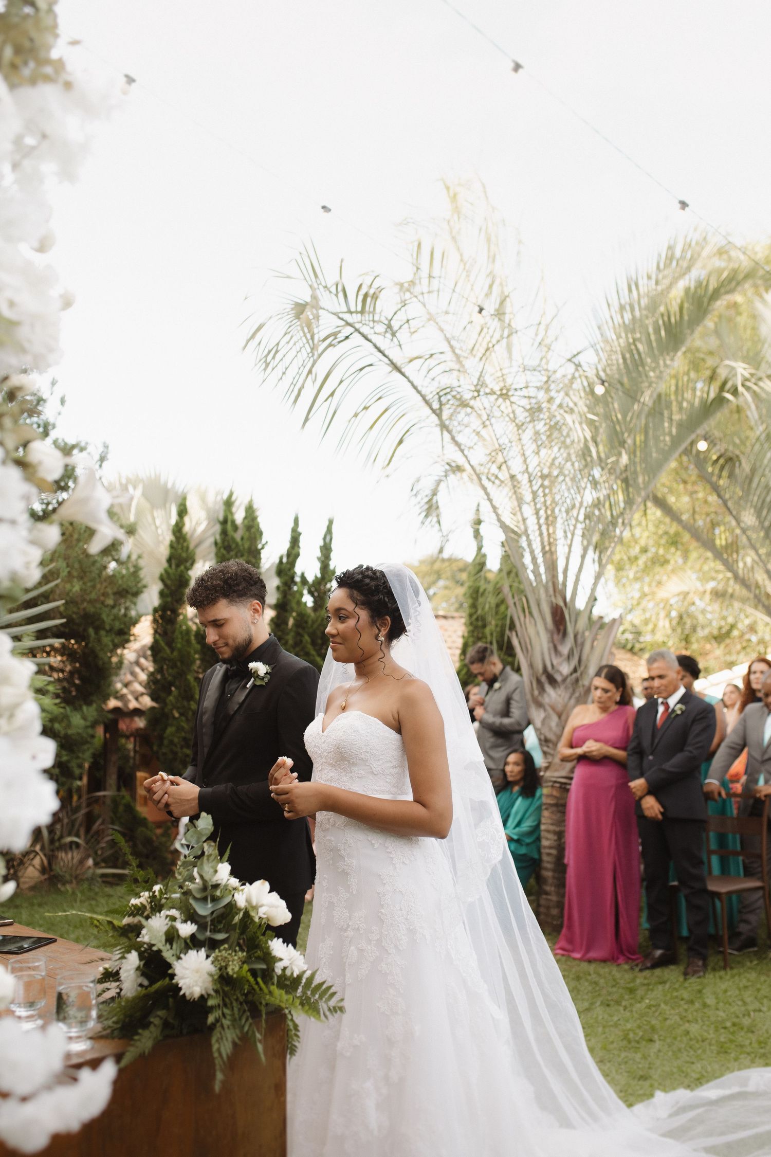 Wedding ceremony taking place outdoors with palm trees and white floral arrangements.