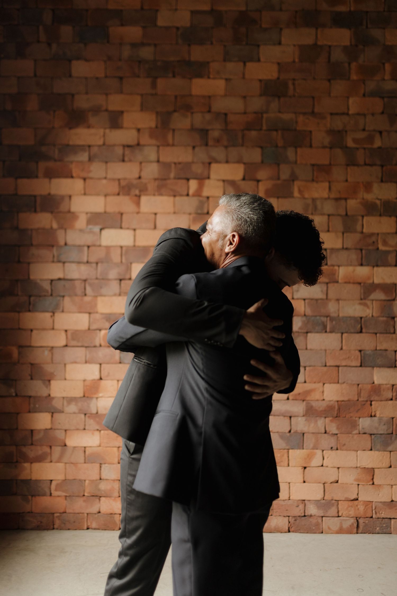 Two people in dark suits share an emotional embrace against a brick wall backdrop.
