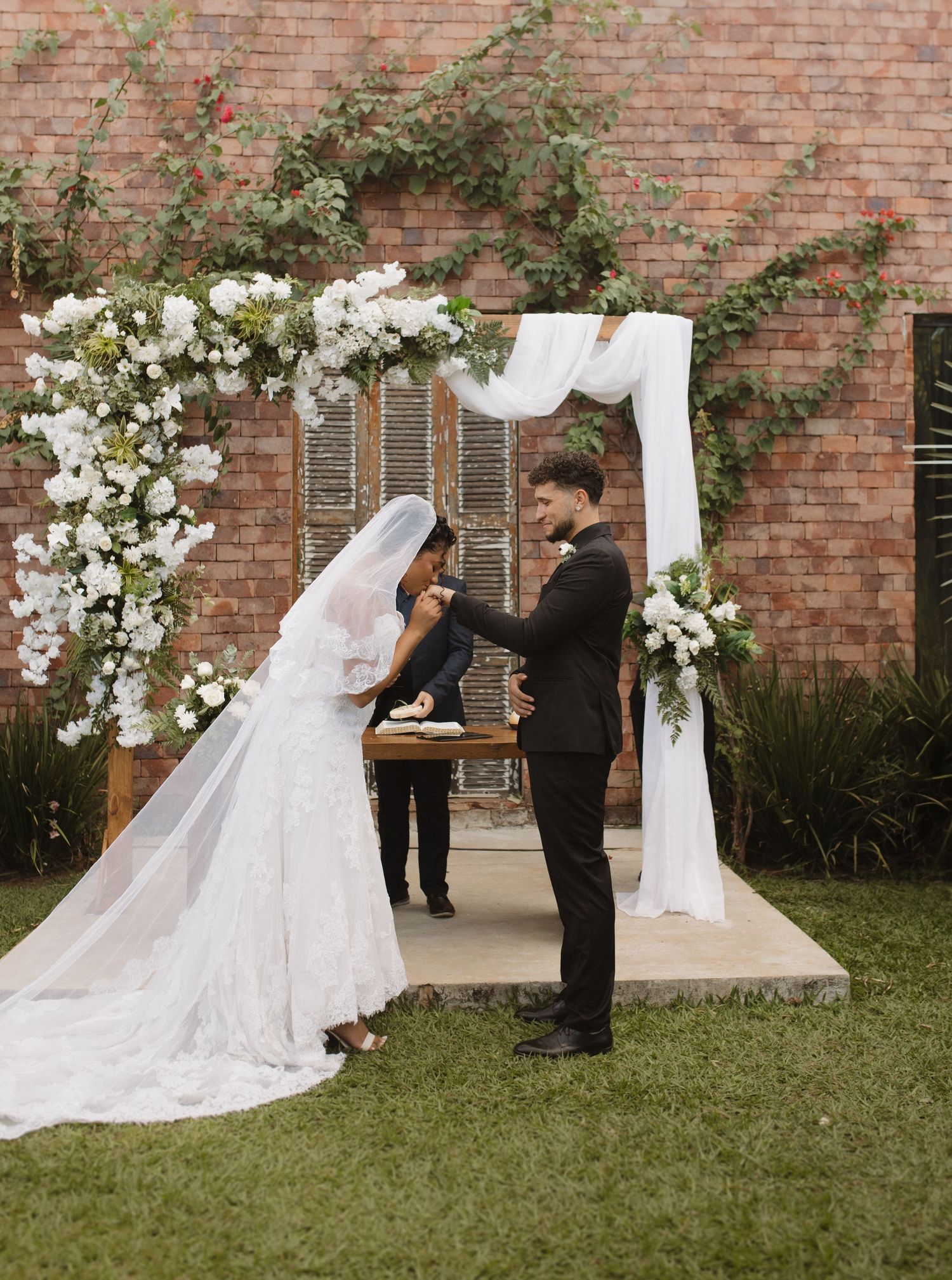 A wedding ceremony in progress with white floral arch and brick wall backdrop.