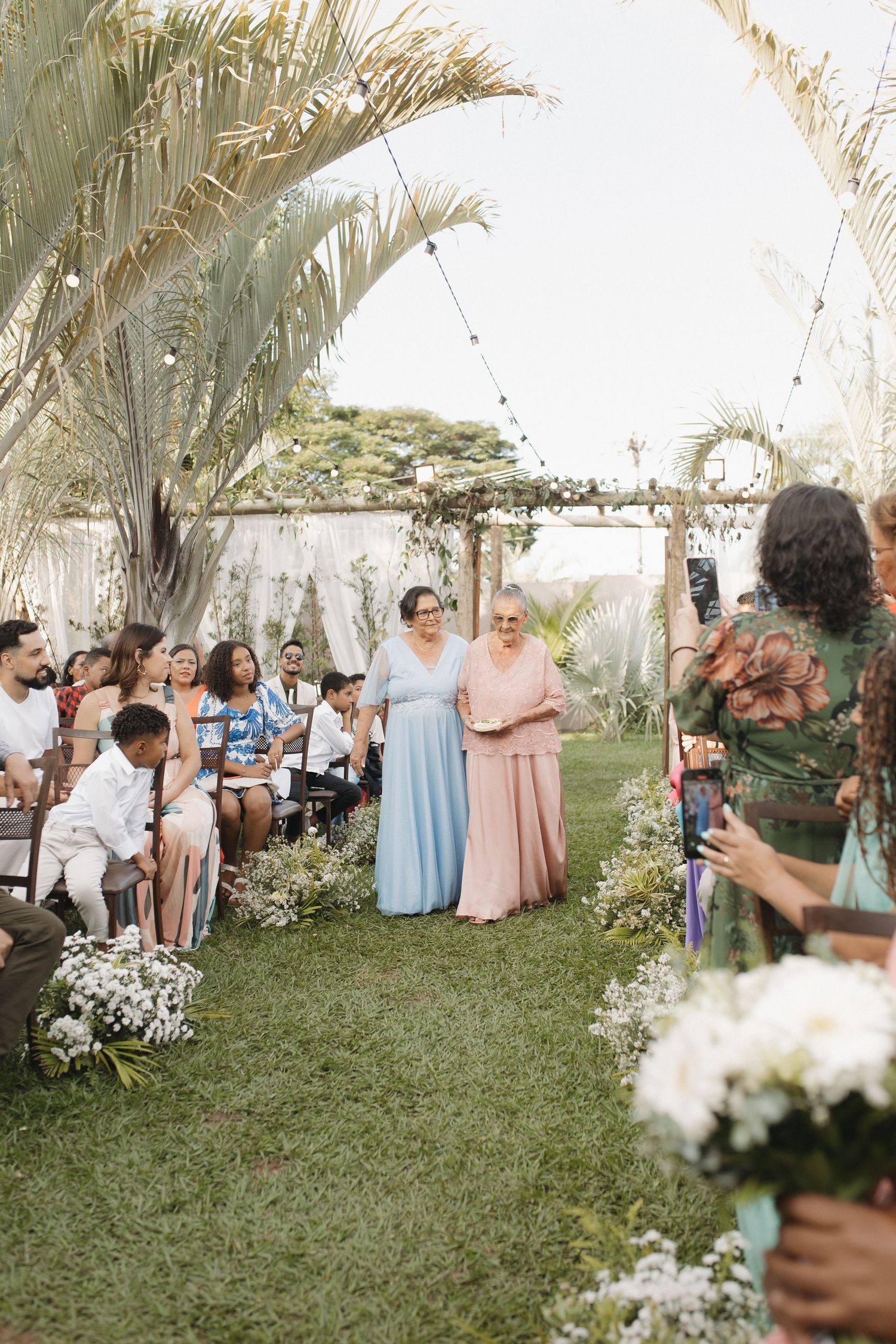Wedding party and guests mingling in a garden setting with palm trees and floral arrangements.