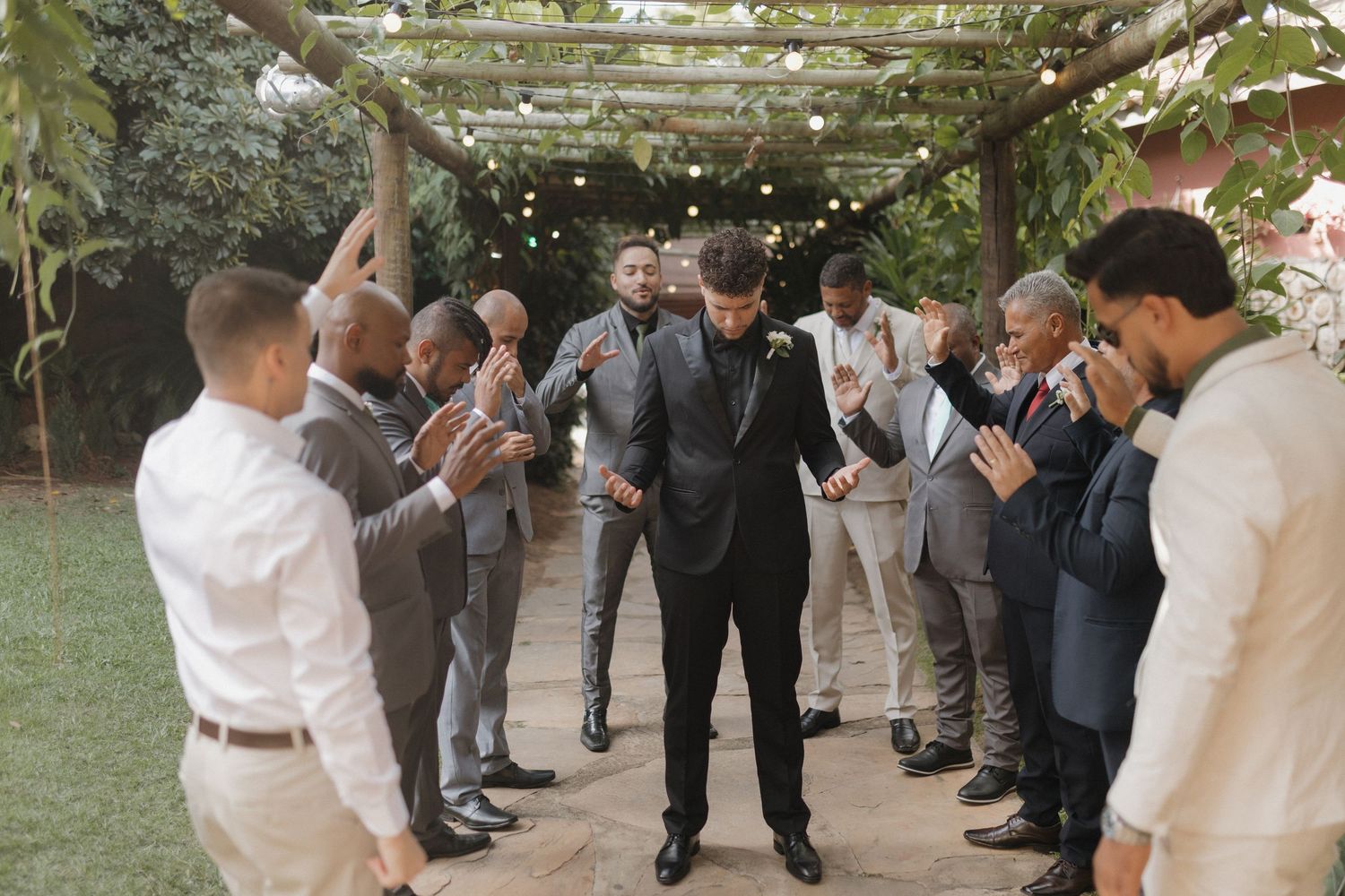 Groomsmen in formal attire stand in a line outdoors during wedding ceremony.