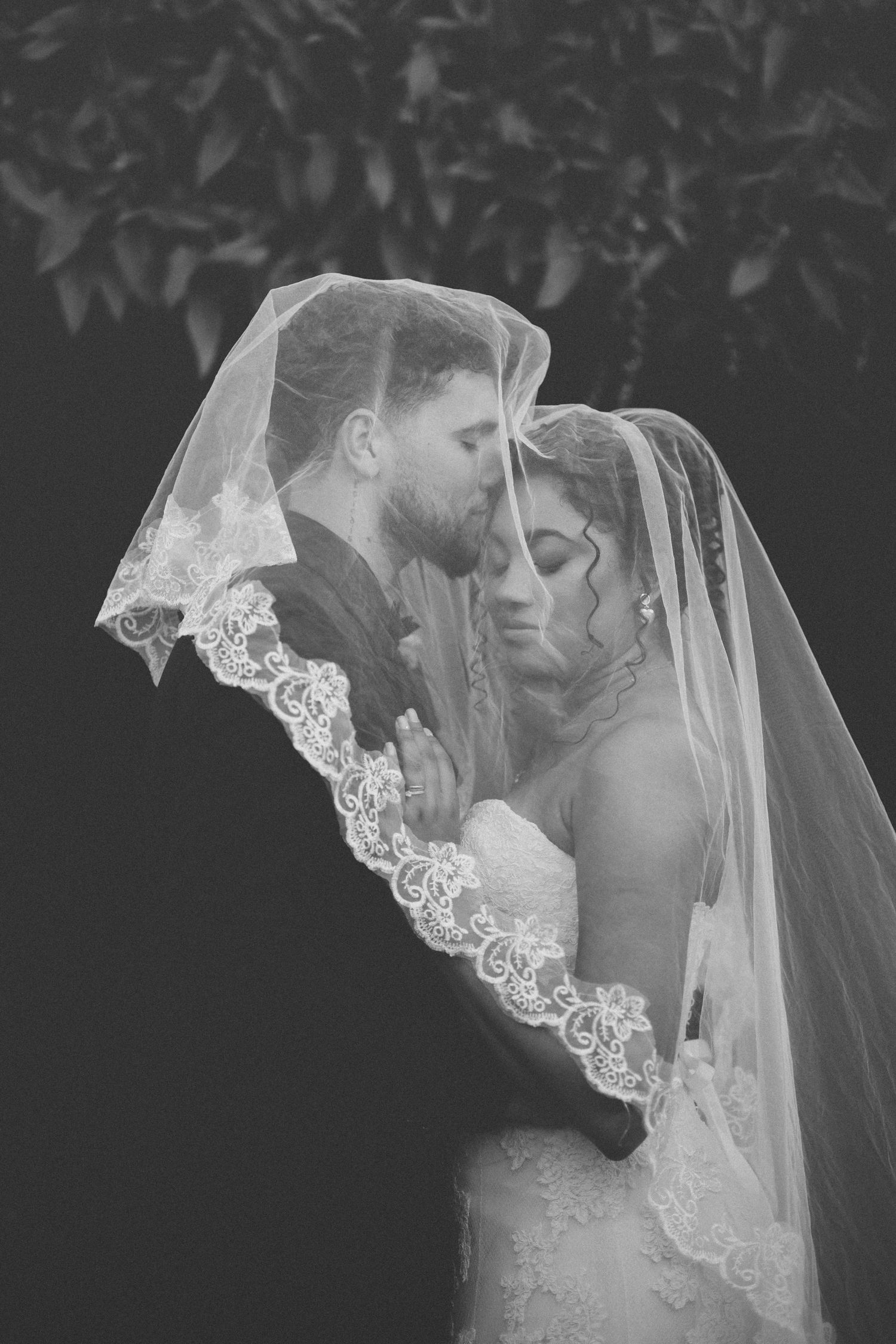 Black and white wedding photo with couple sharing romantic moment under lace veil.