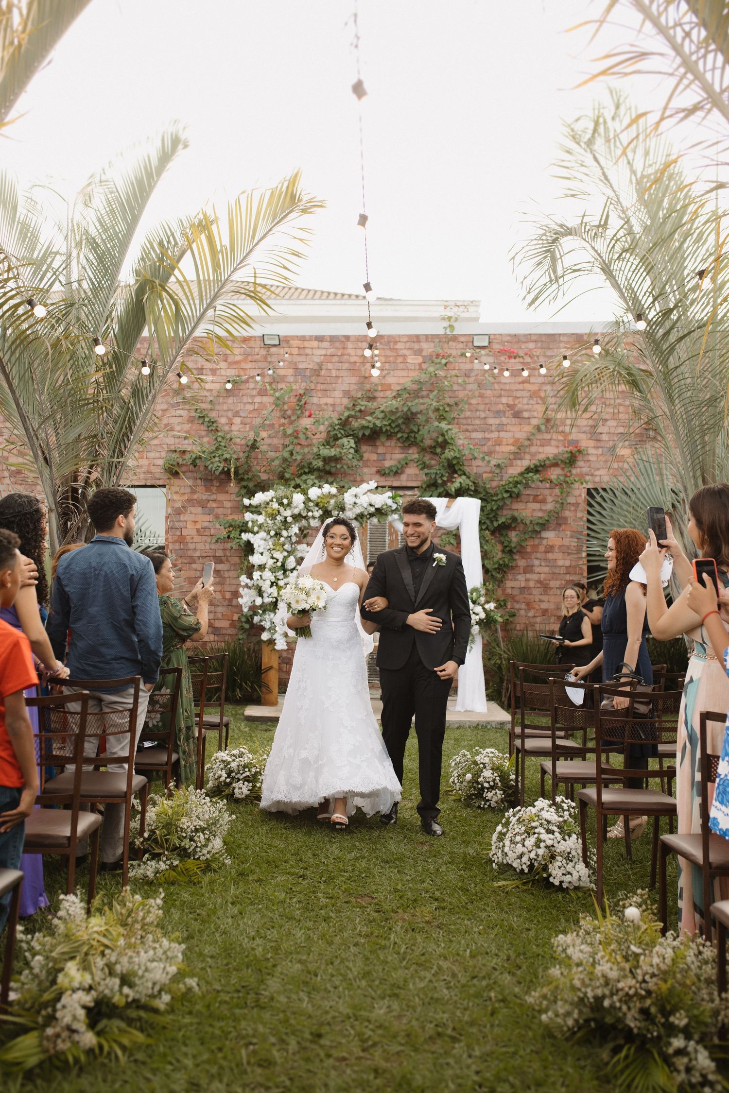Wedding guests gather in a garden courtyard with palm trees and brick walls.