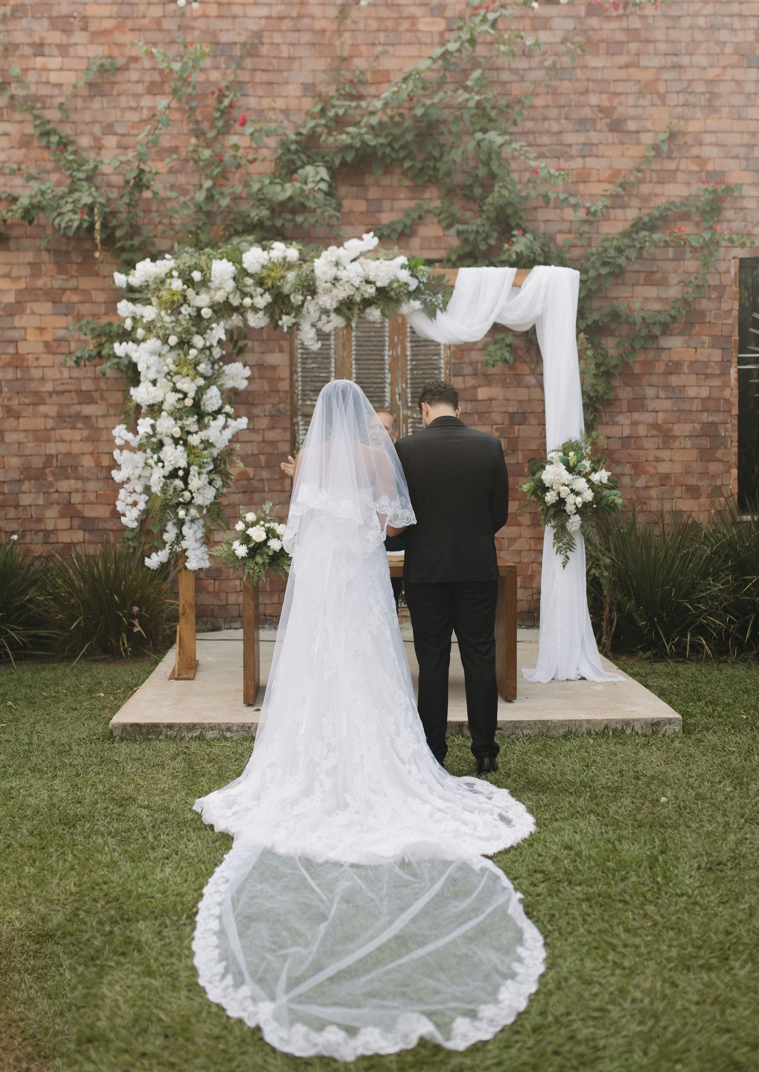 Wedding couple photographed from behind against brick wall with white floral arrangements.