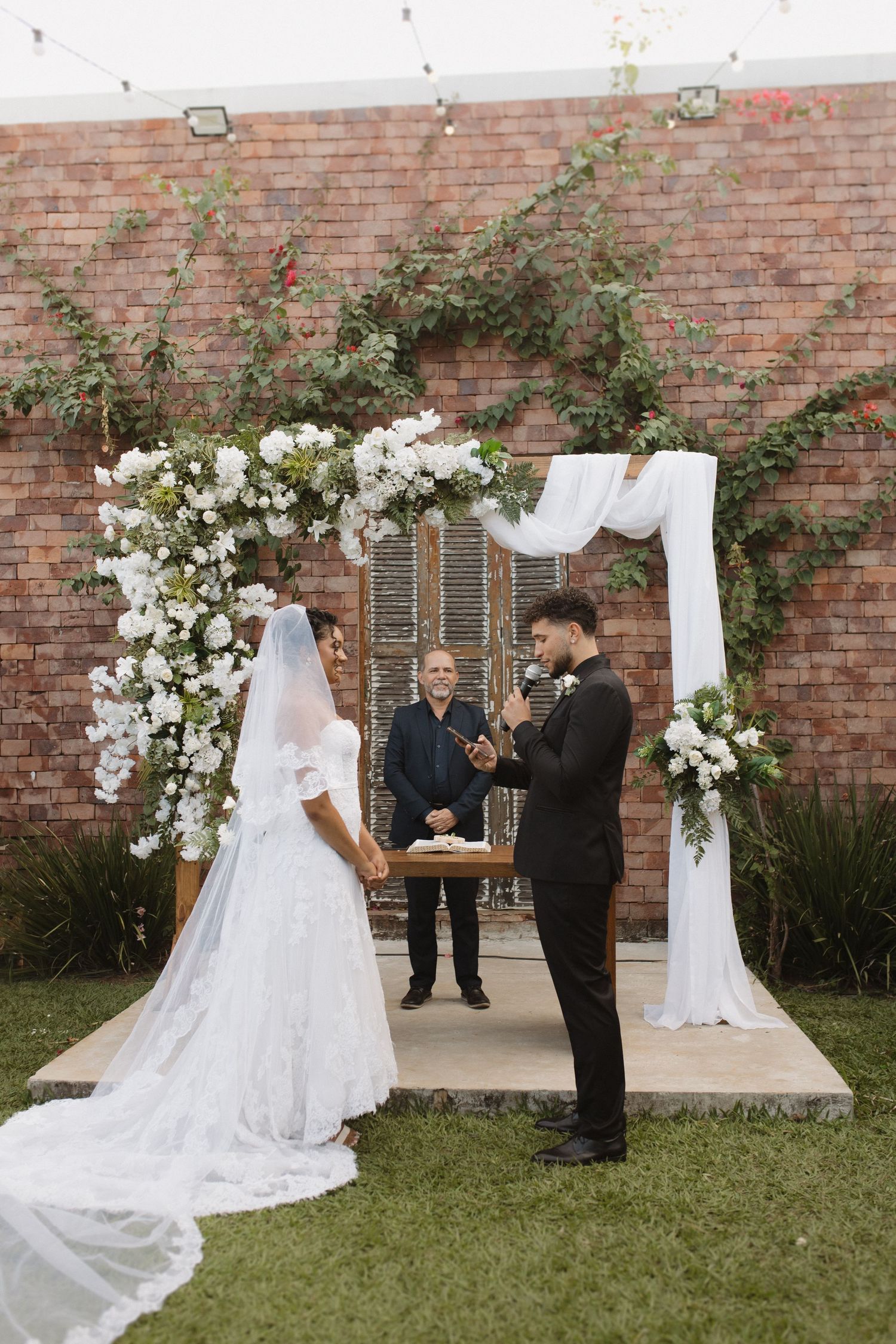 An outdoor wedding ceremony setup with a floral arch against a brick wall and white decorations.