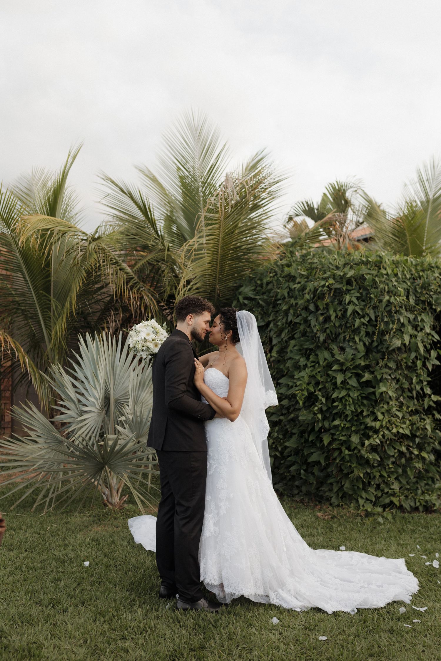 Romantic wedding couple embraces in a tropical garden setting with palm trees and greenery.