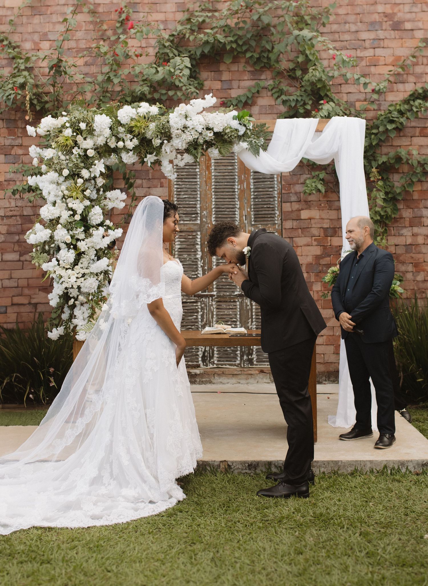 A wedding ceremony scene with white floral arrangements against a rustic brick backdrop.