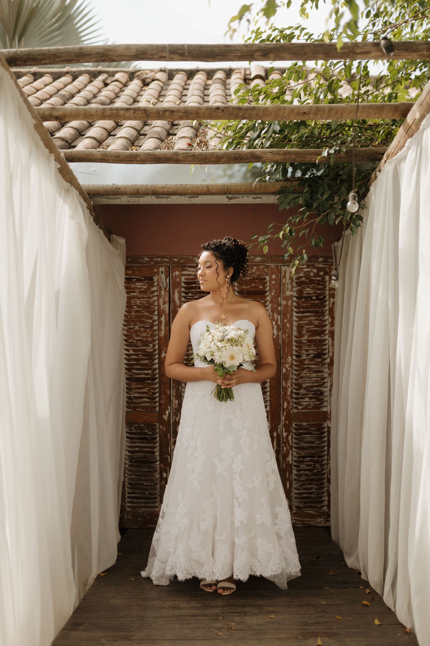 Bride in a beautiful white gown poses in front of a rustic wall with flowing white curtains.
