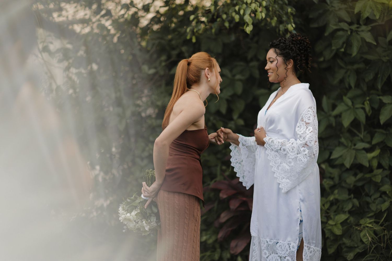 Two people share a tender moment outdoors, one wearing white lace and the other in brown.