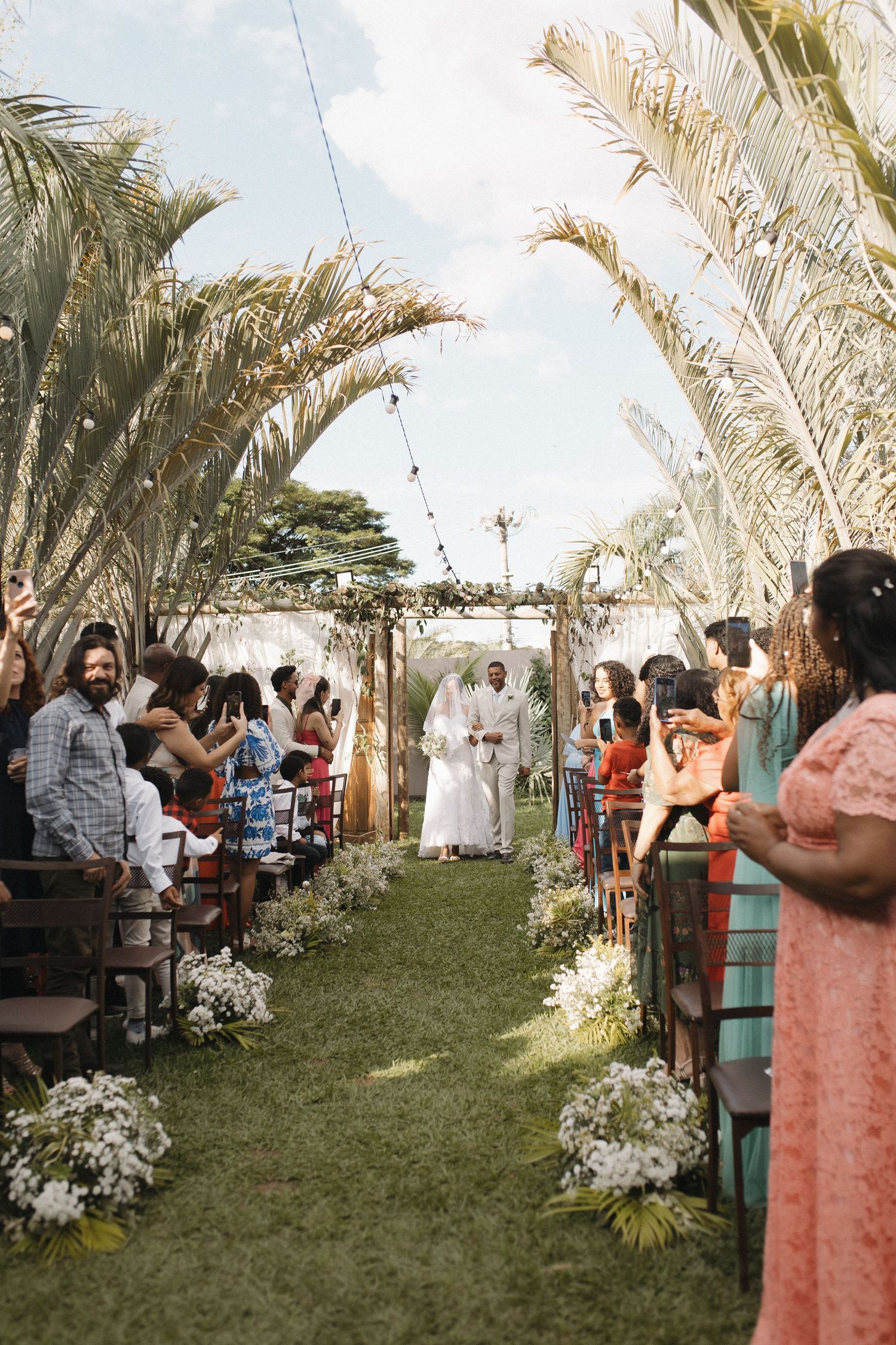 Wedding ceremony setup with palm trees and white decor in outdoor garden setting.