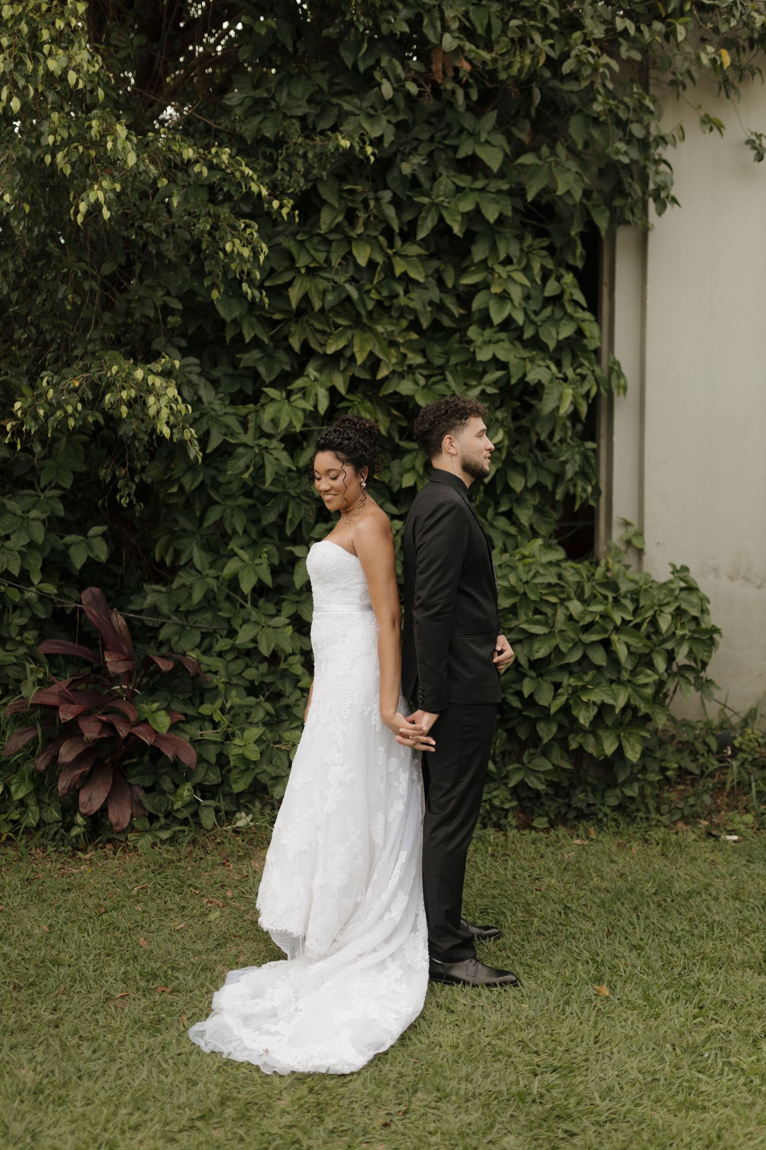 Wedding couple poses against a wall covered in lush green foliage.