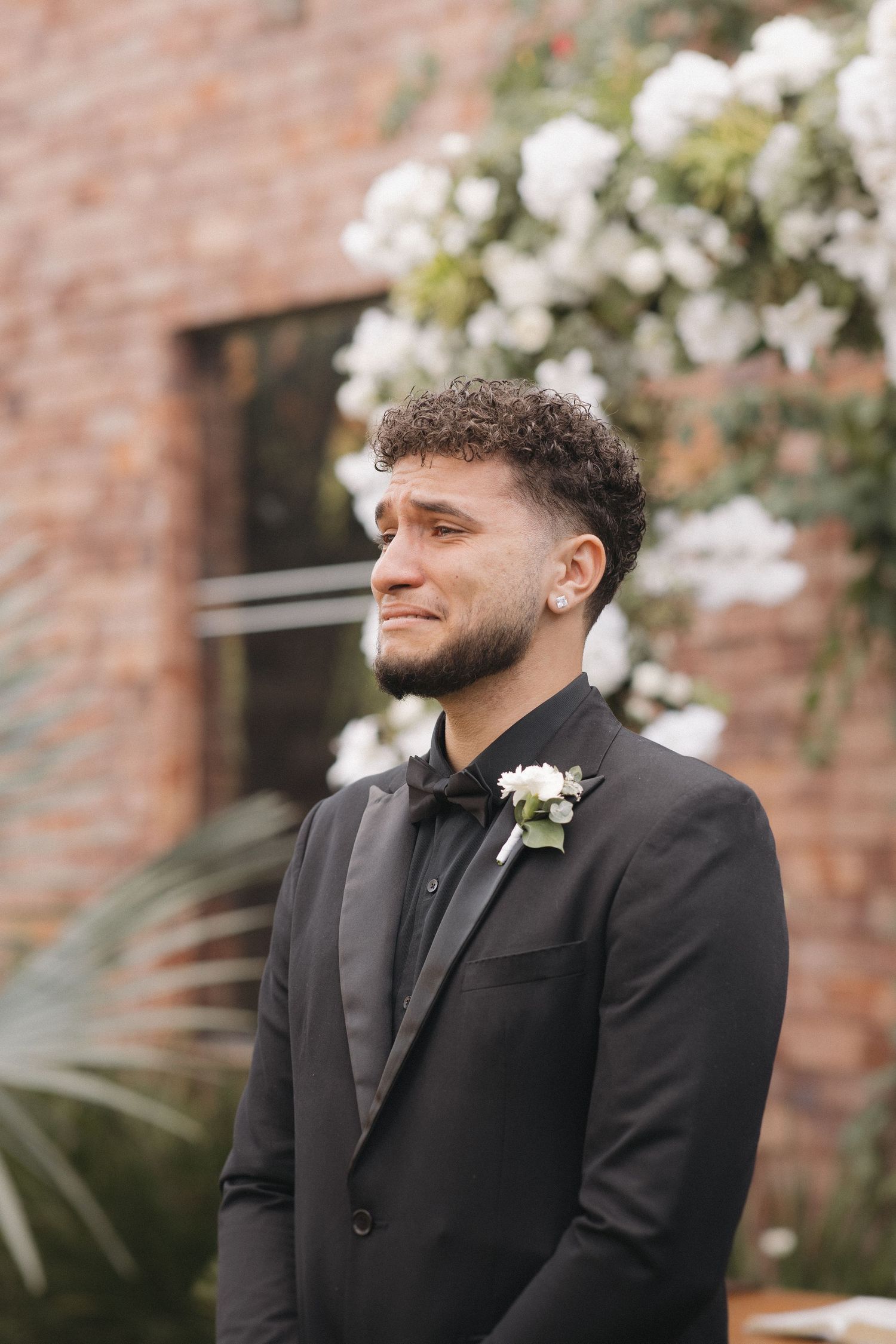 Groom in black suit with boutonniere stands at outdoor wedding venue with brick backdrop.
