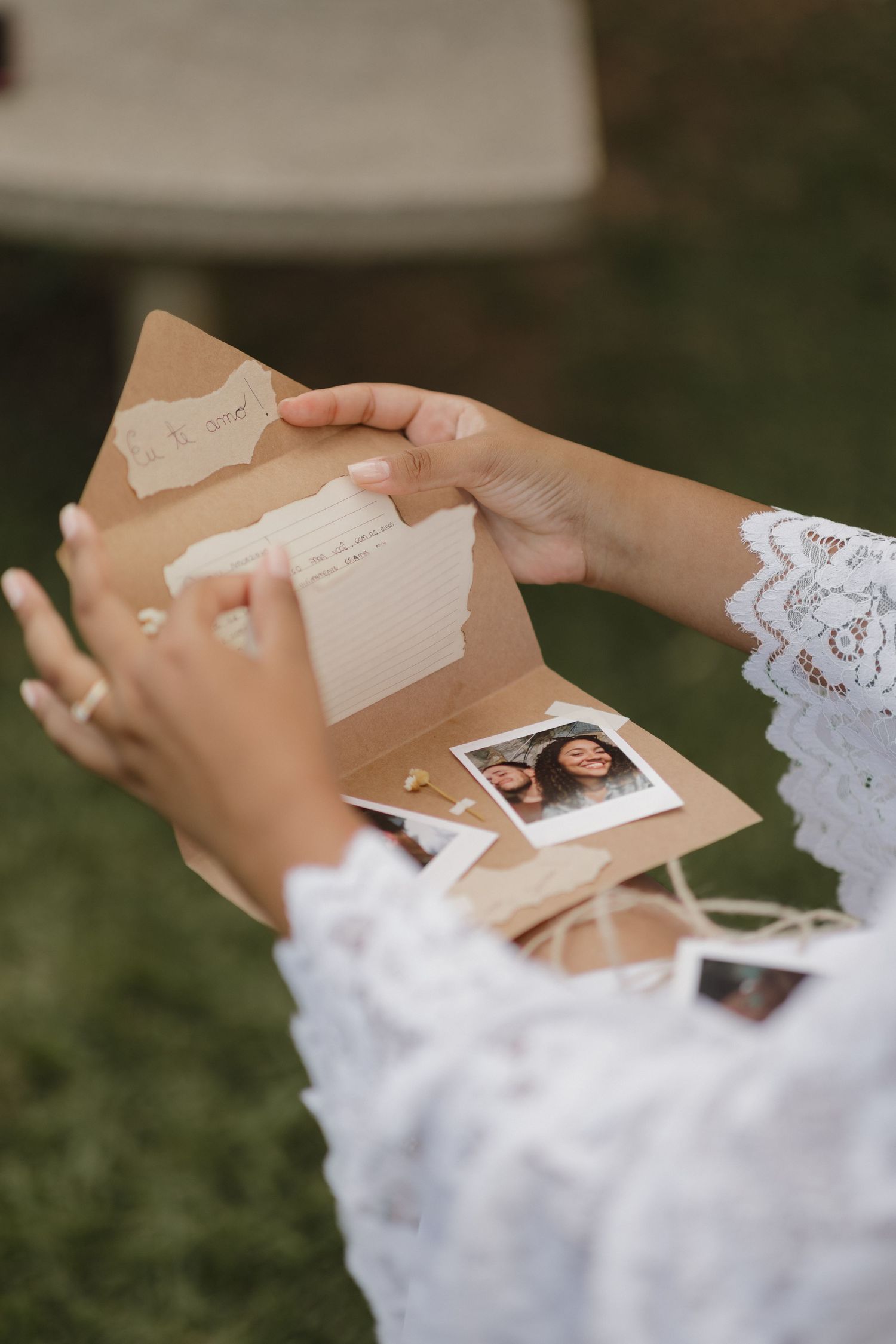 Hands in white lace sleeves hold photographs during an outdoor wedding preparation session.