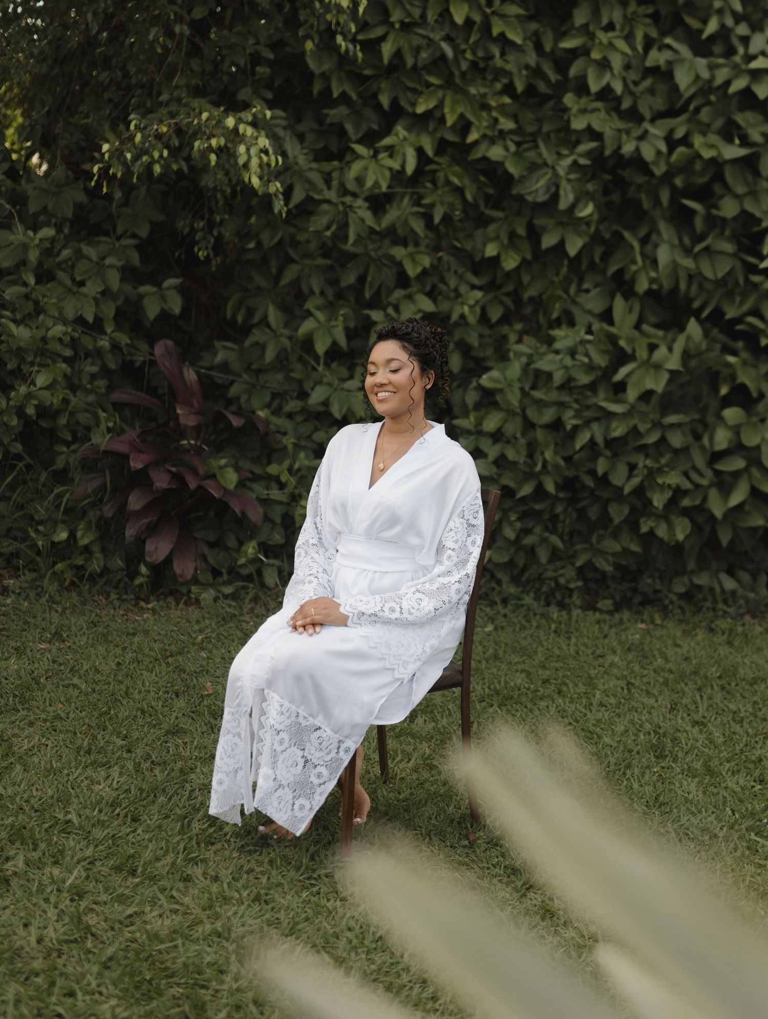 A person in a white lace robe sits gracefully in a garden setting with lush green foliage.