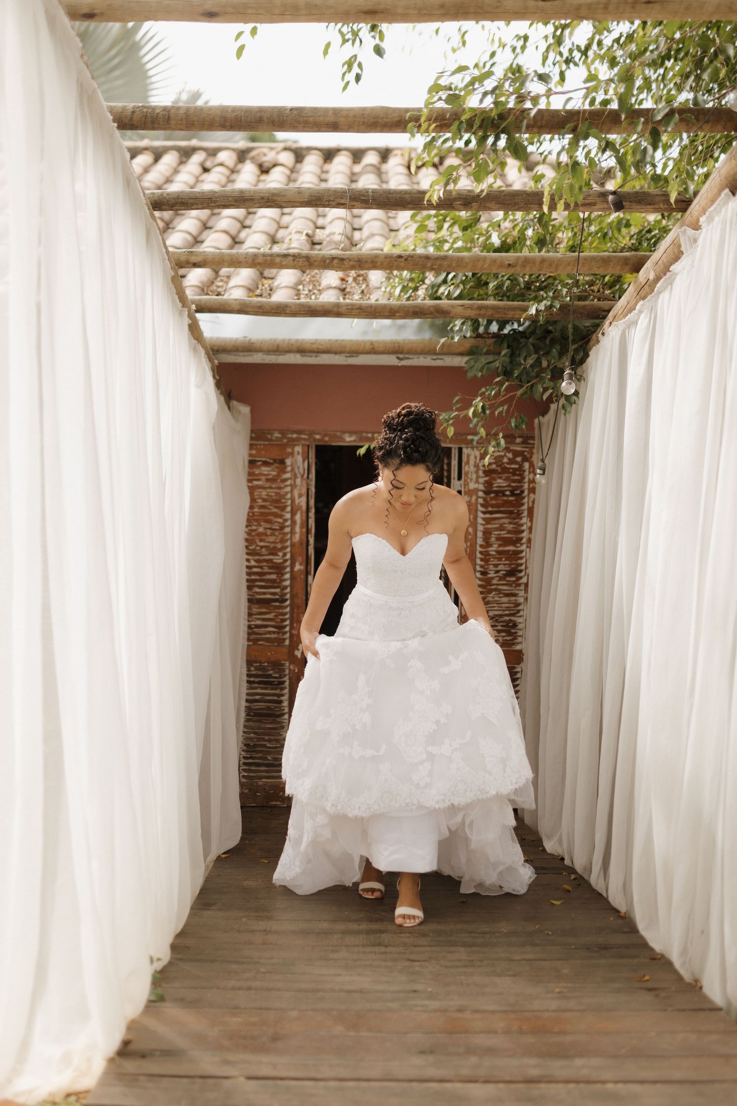 A person in a flowing white wedding gown sits gracefully against white curtains in an outdoor setting.