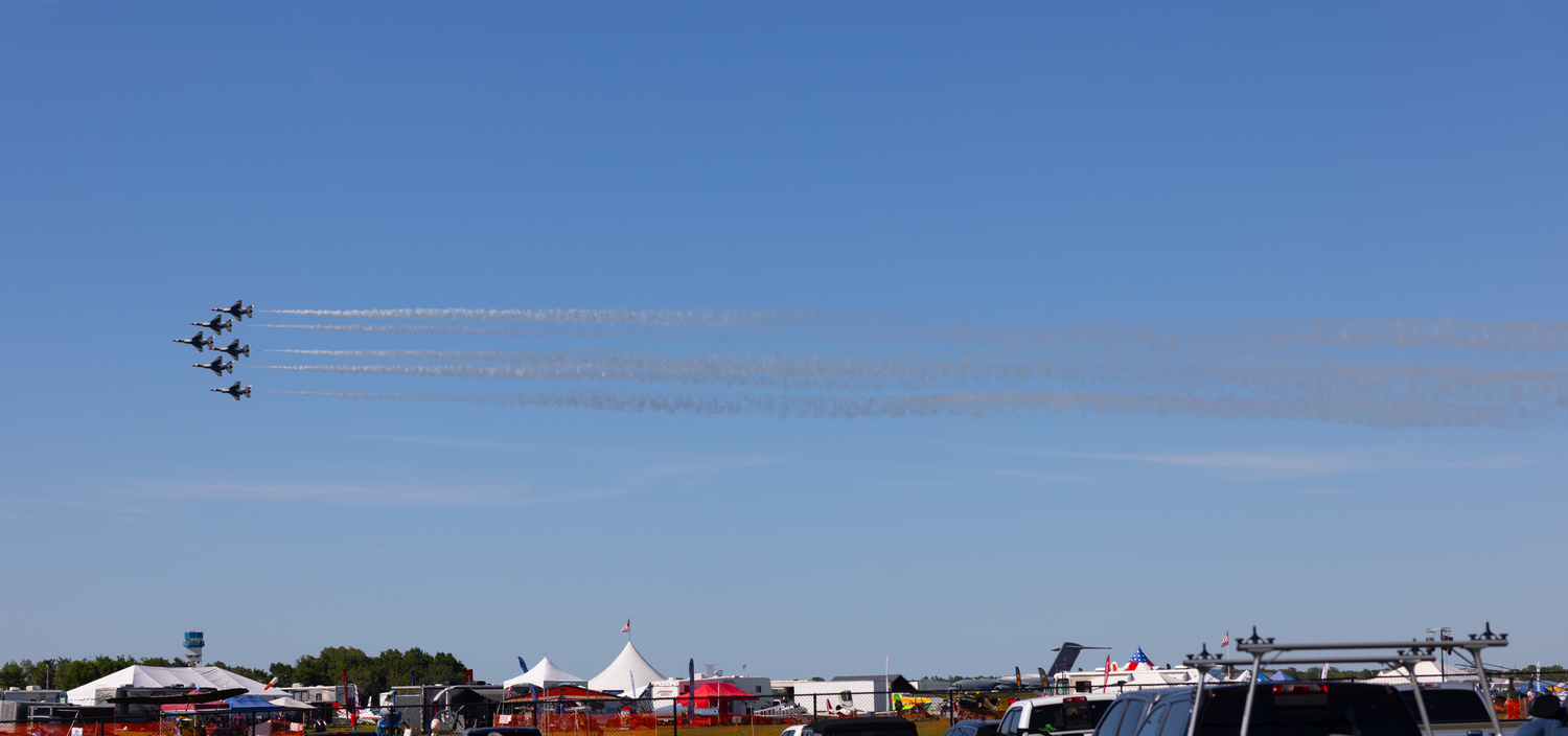 Military jets flying in tight formation with smoke trails over a crowded airshow grounds.