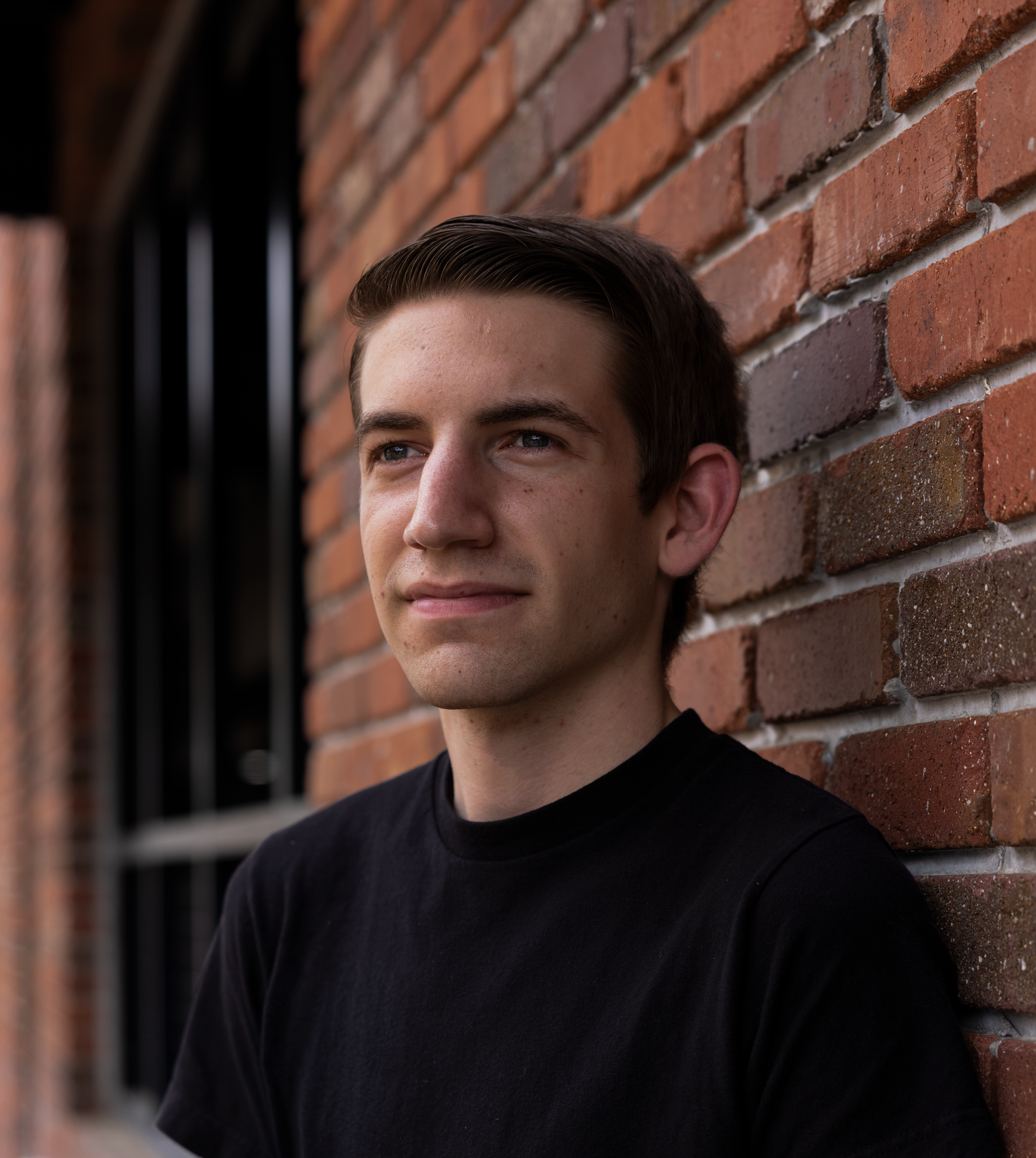 Young man in black t-shirt leaning against a red brick wall outdoors.