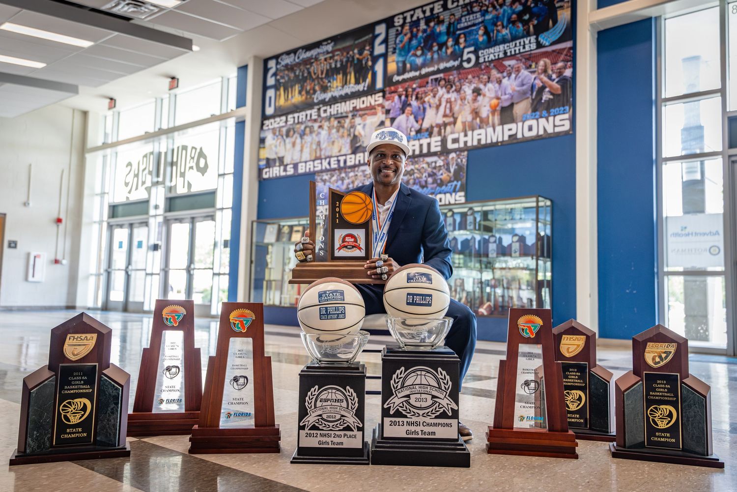 Man posing with multiple basketball championship trophies in a school trophy display area.