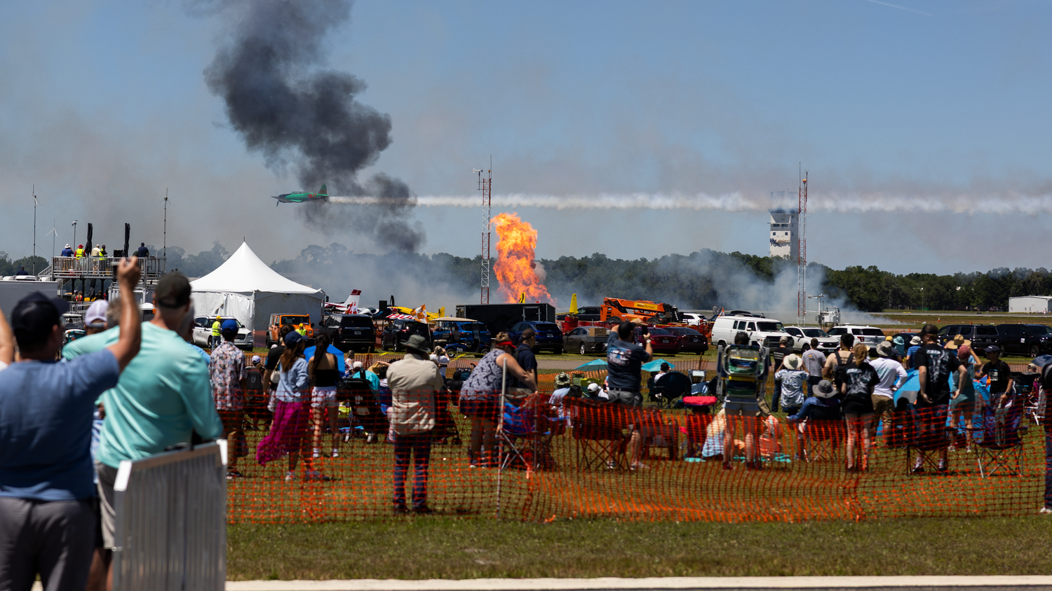 Airshow crowd watches a dramatic fire explosion display on the tarmac with smoke rising into the sky.