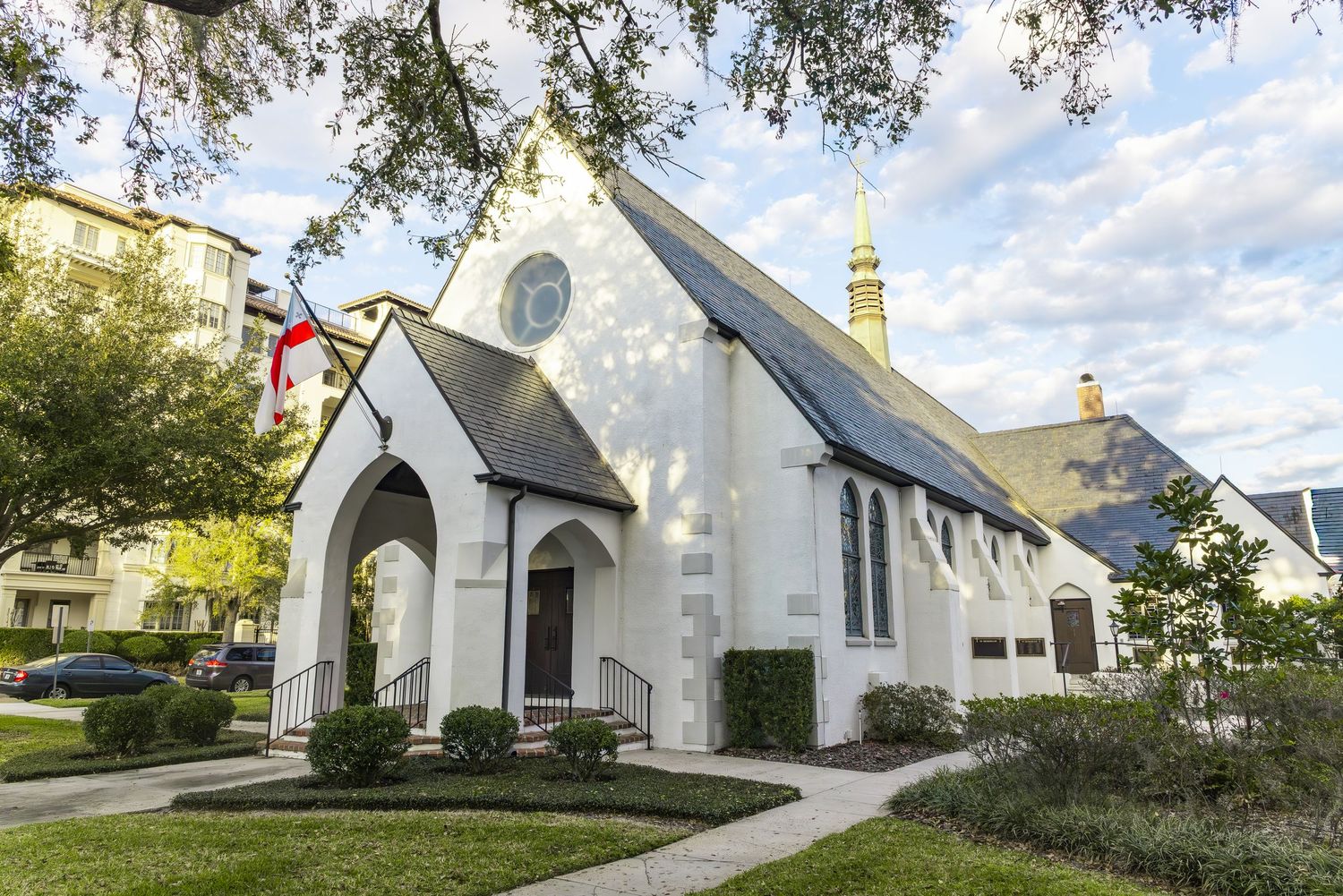 White historic church with Gothic arched entrance surrounded by green trees and landscaping.