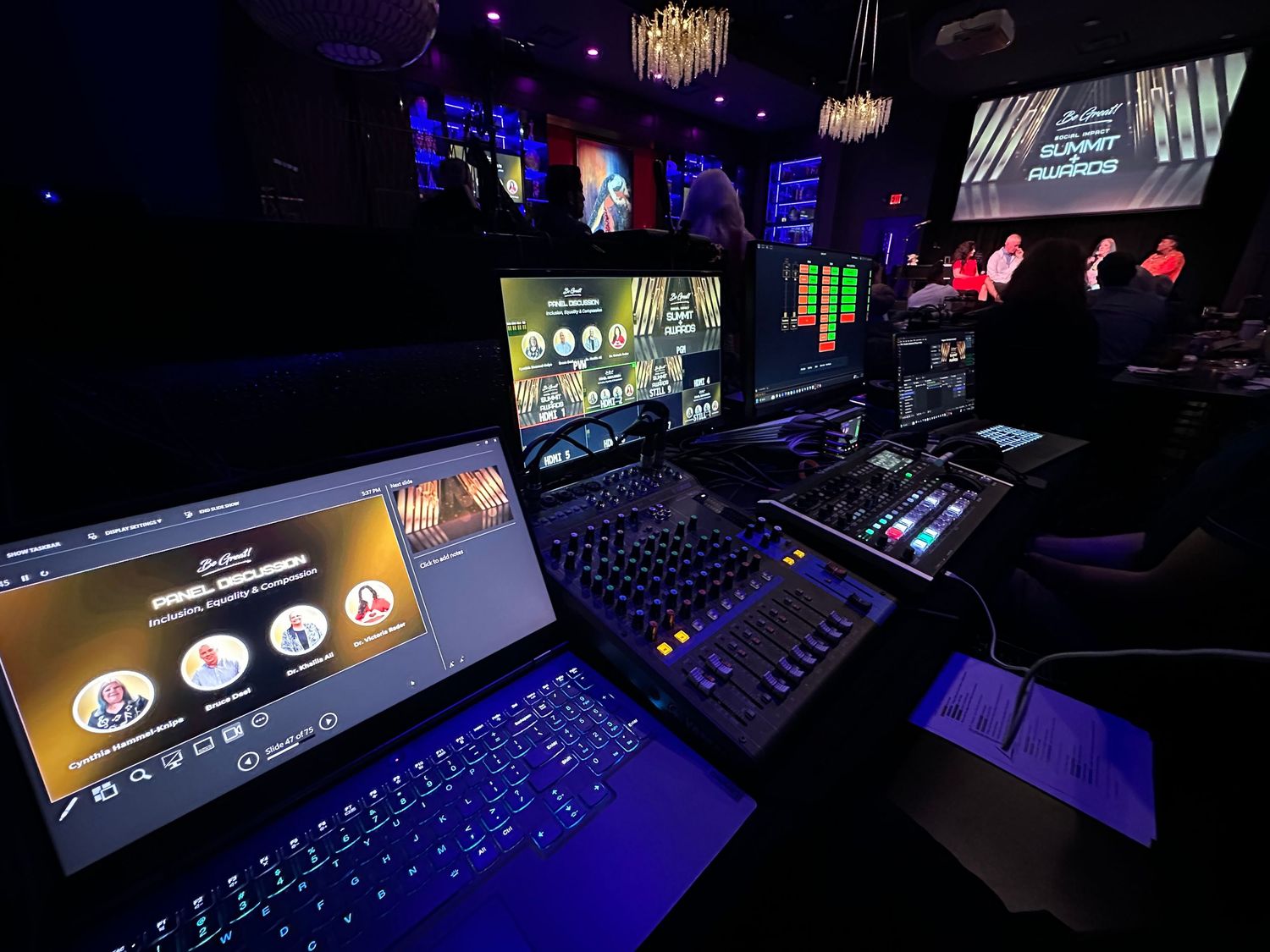 Live event production setup with laptops, mixing board, and multiple screens in a dark venue.
