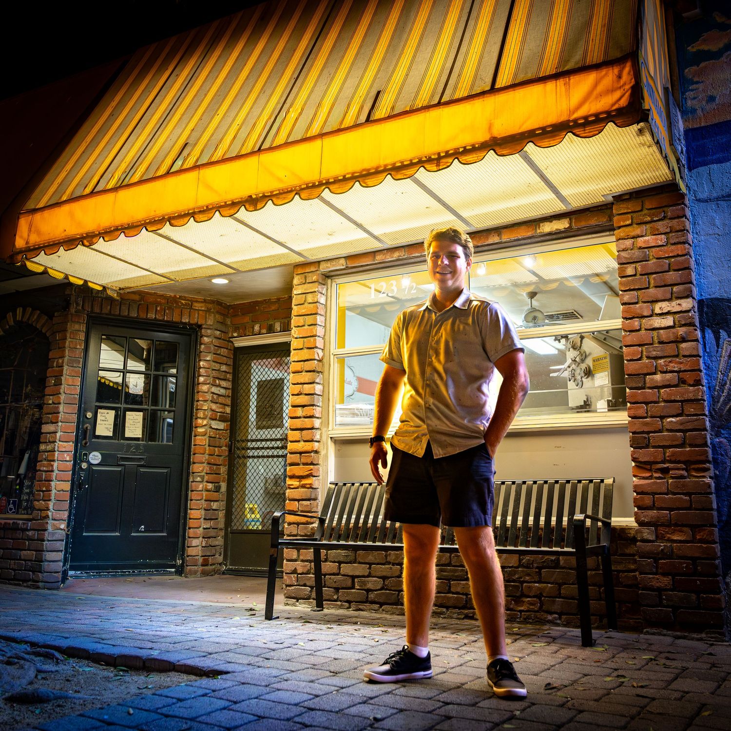 Young man standing outside a lit storefront at night wearing a yellow shirt and shorts.