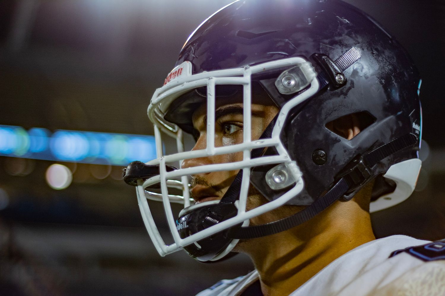 Close-up of a football player wearing a dark helmet with face mask under stadium lights.
