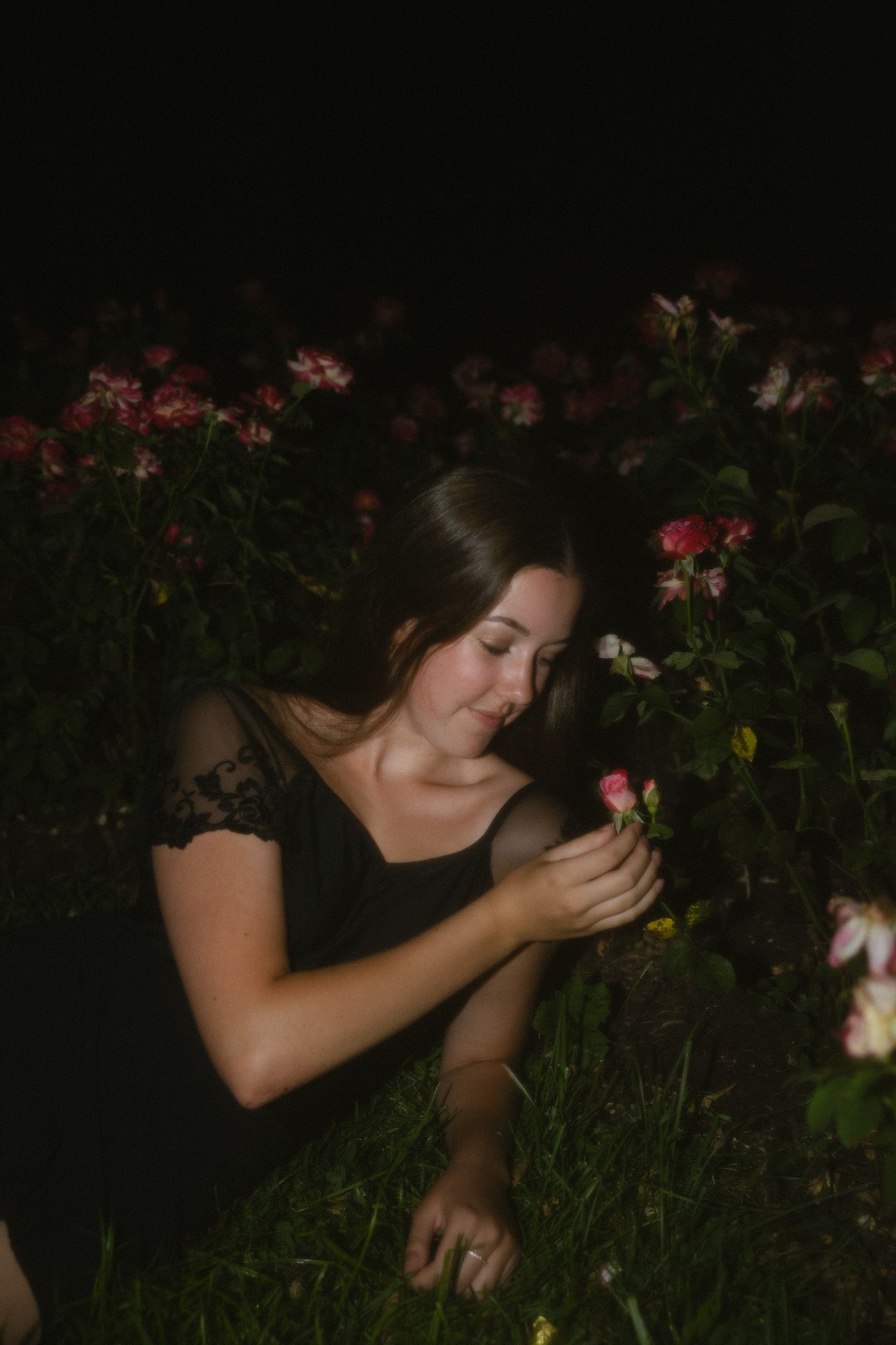 A young woman in a black lace dress kneels among blooming pink roses at night, gently holding a flower.