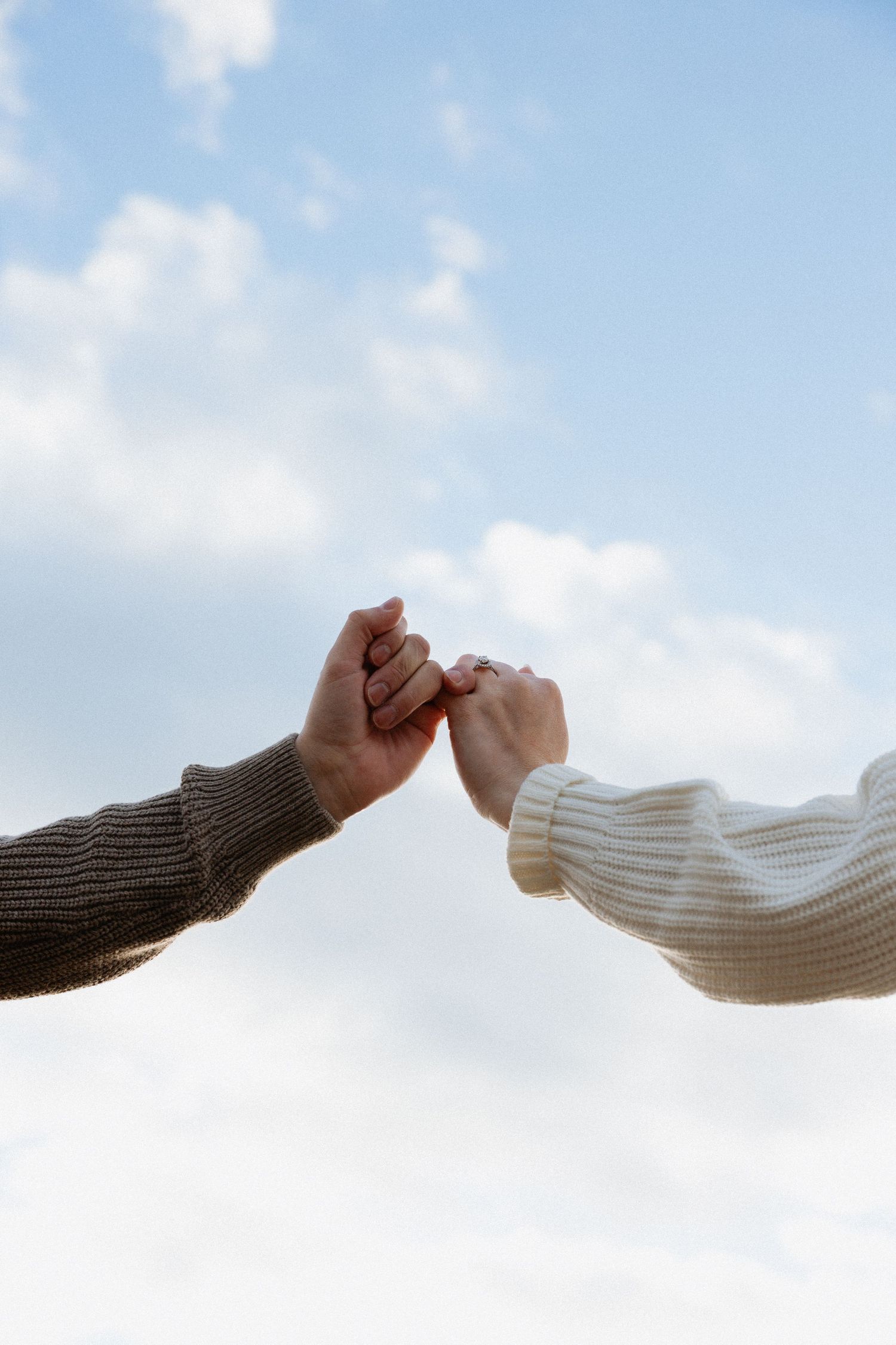 Two hands in sweater sleeves reach toward each other against a cloudy sky background in a gesture of connection.