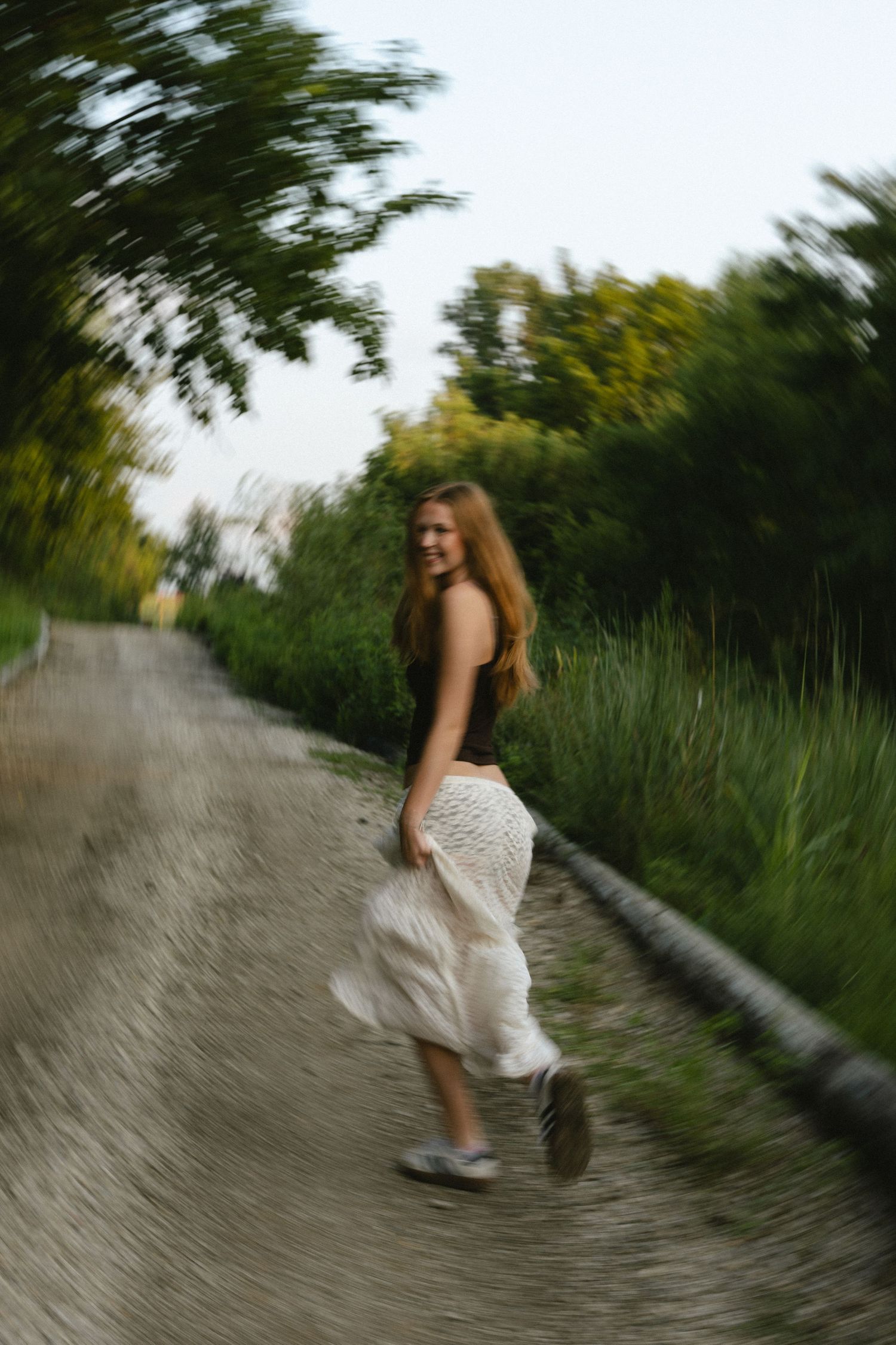 Young woman in white skirt walking on a rural path surrounded by lush green trees and grass.