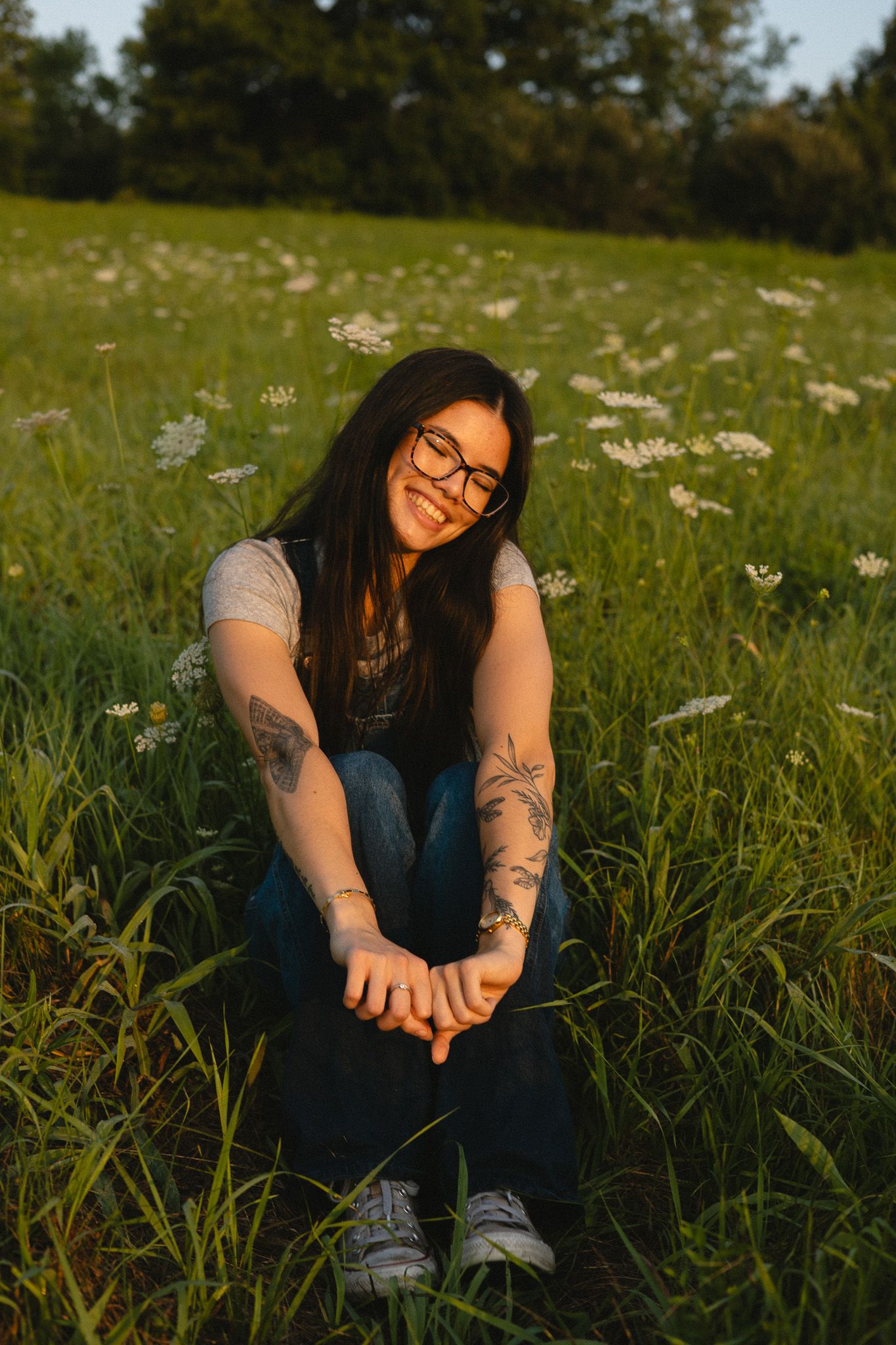 Smiling young woman with tattoos and glasses sitting in a sunny green meadow with wildflowers at golden hour.