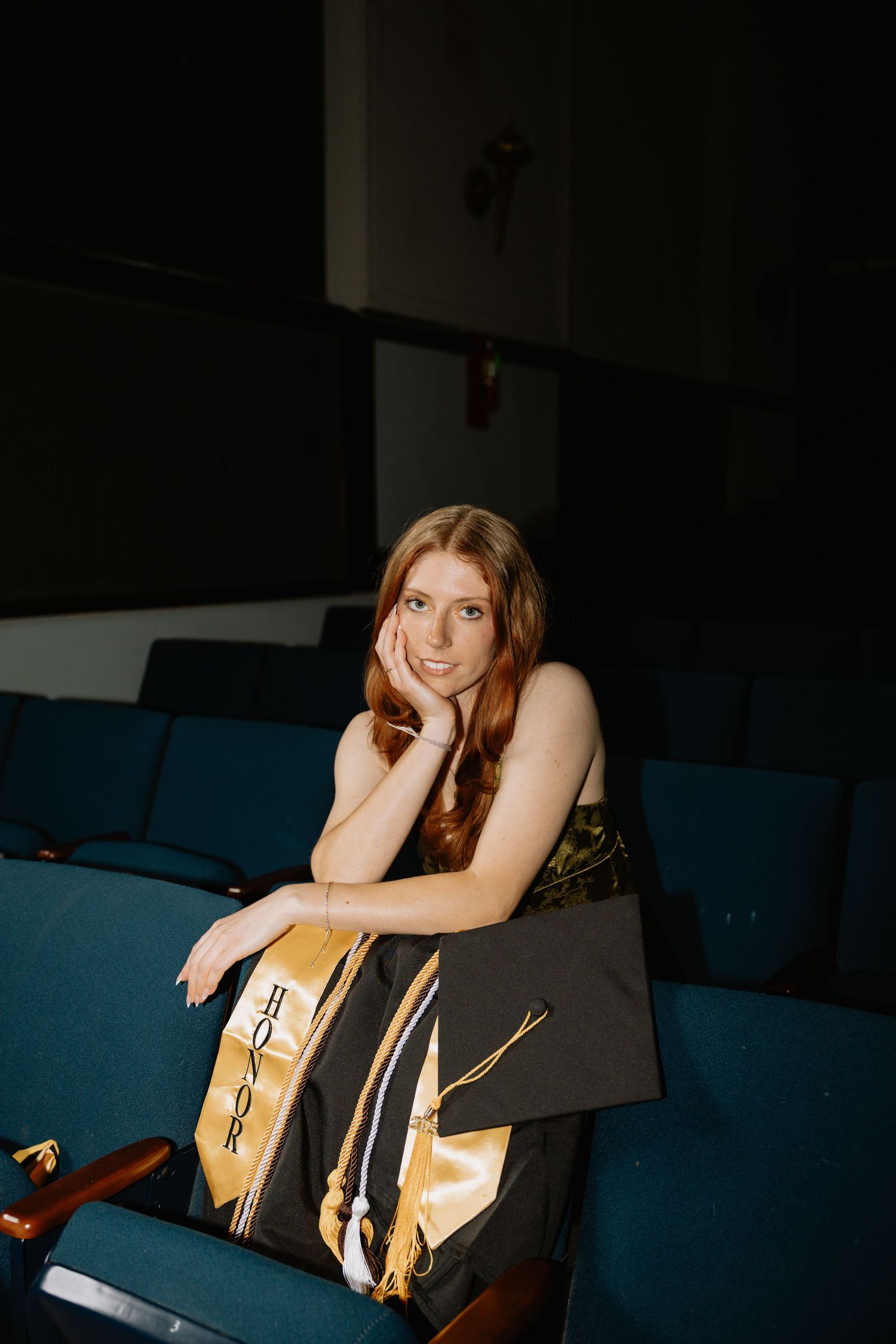 Graduate in dark auditorium wearing gold stole and graduation cap.