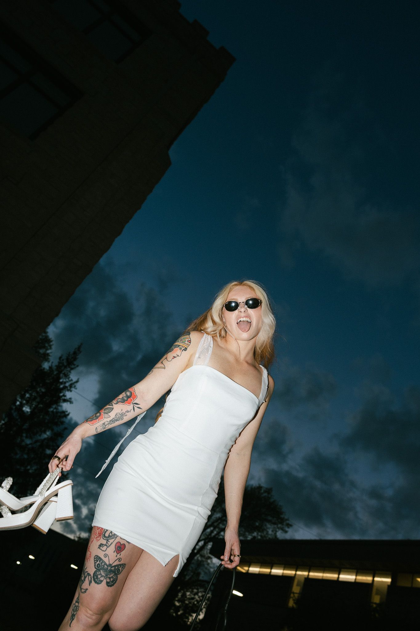 Blonde woman in white mini dress with tattoos, sunglasses, holding heels outdoors at night.
