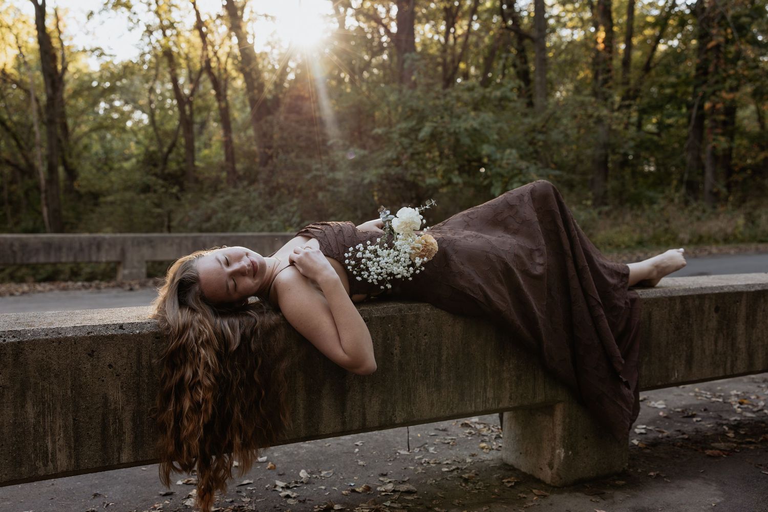A person in a black dress lies gracefully on a stone wall at sunset in a wooded area.