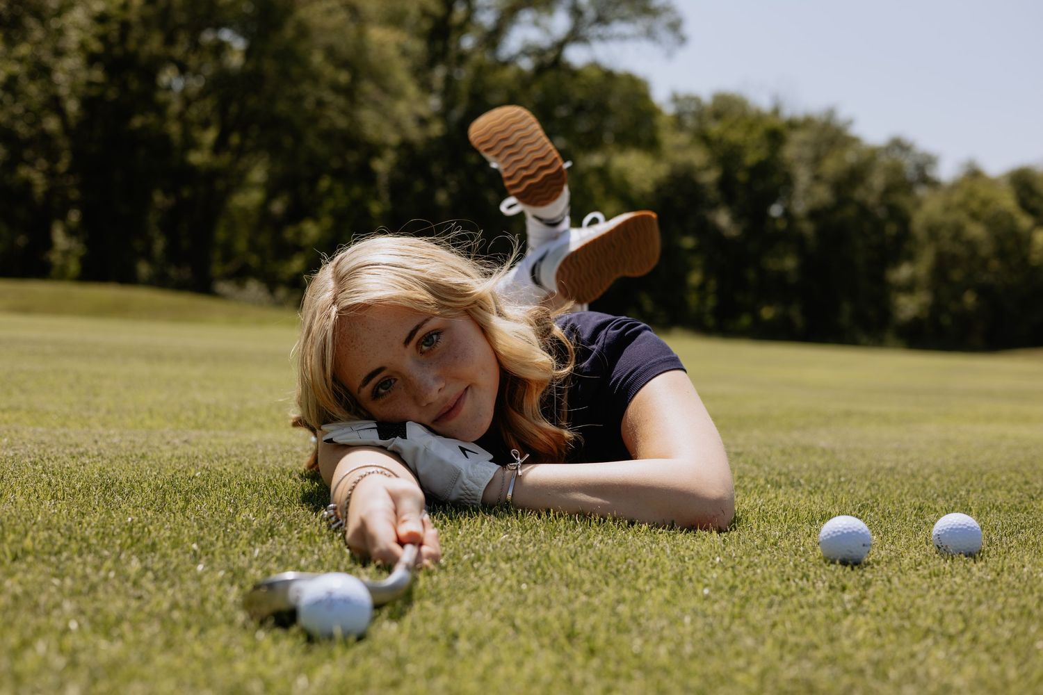 Golfer lying on grass with multiple golf balls scattered on putting green during sunny day.