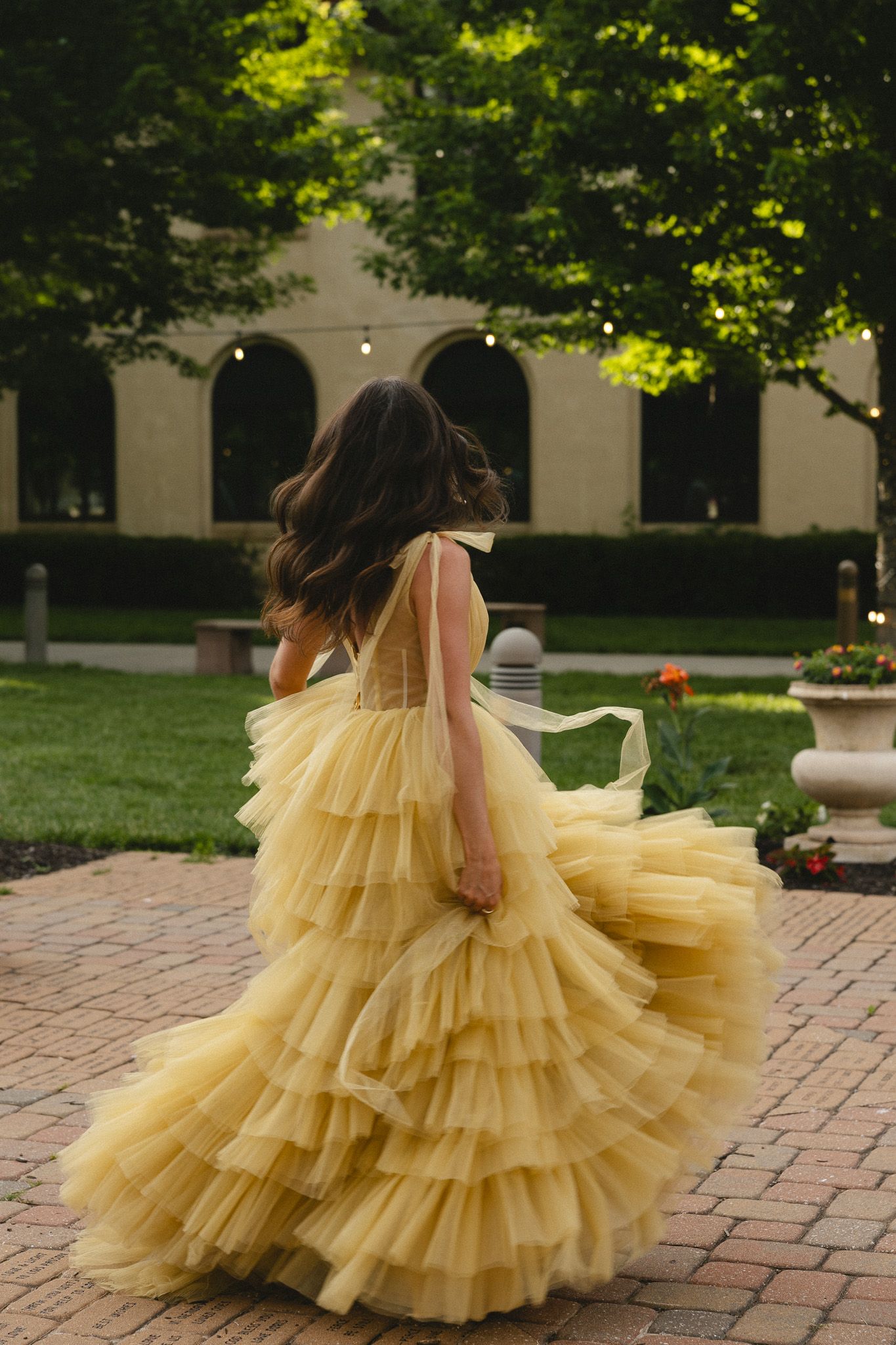 Woman in yellow ruffled tulle ball gown twirling on brick path in lush garden setting.