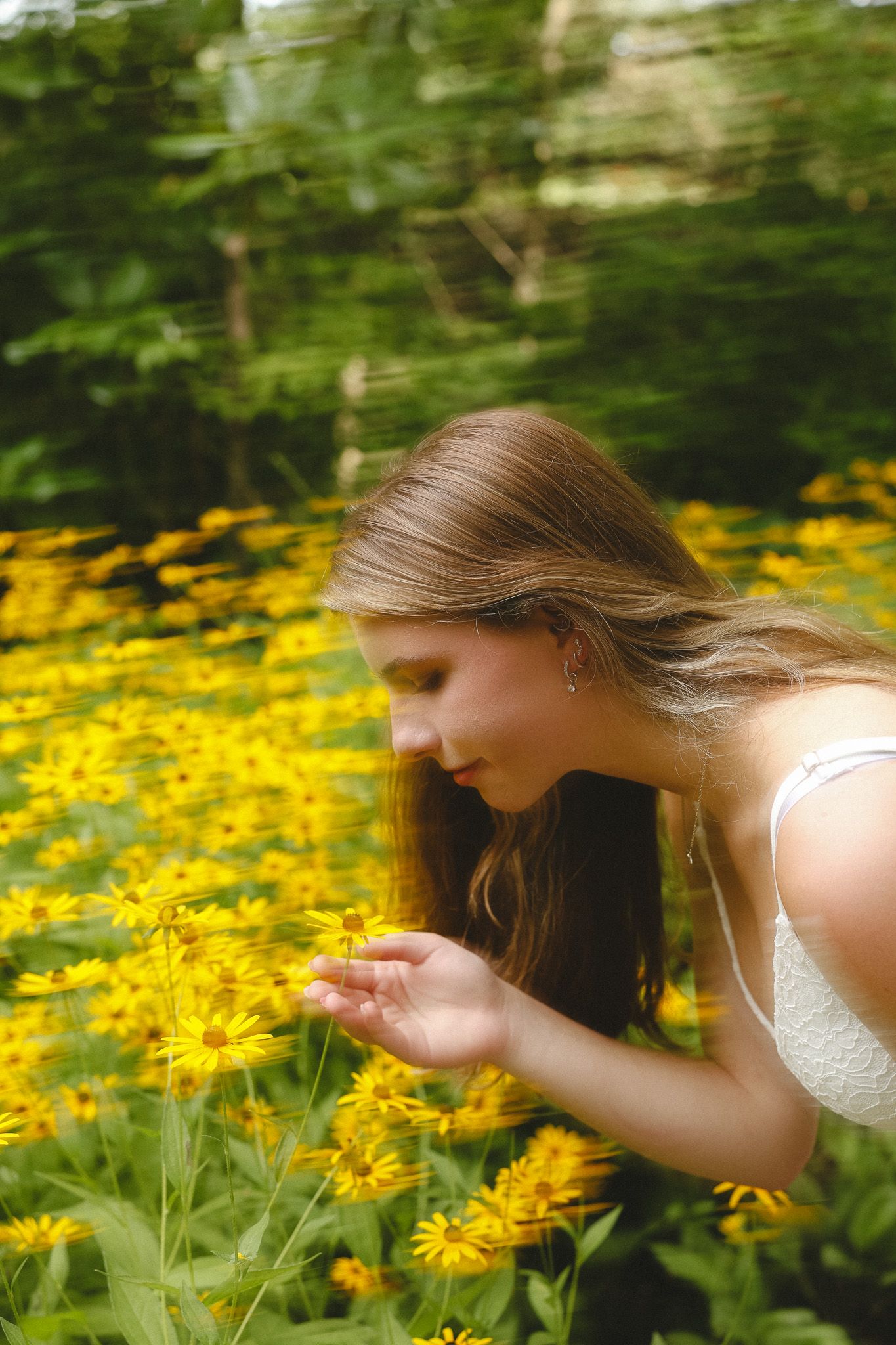 Young woman with long hair smelling yellow wildflowers in a lush green meadow, wearing a white dress.