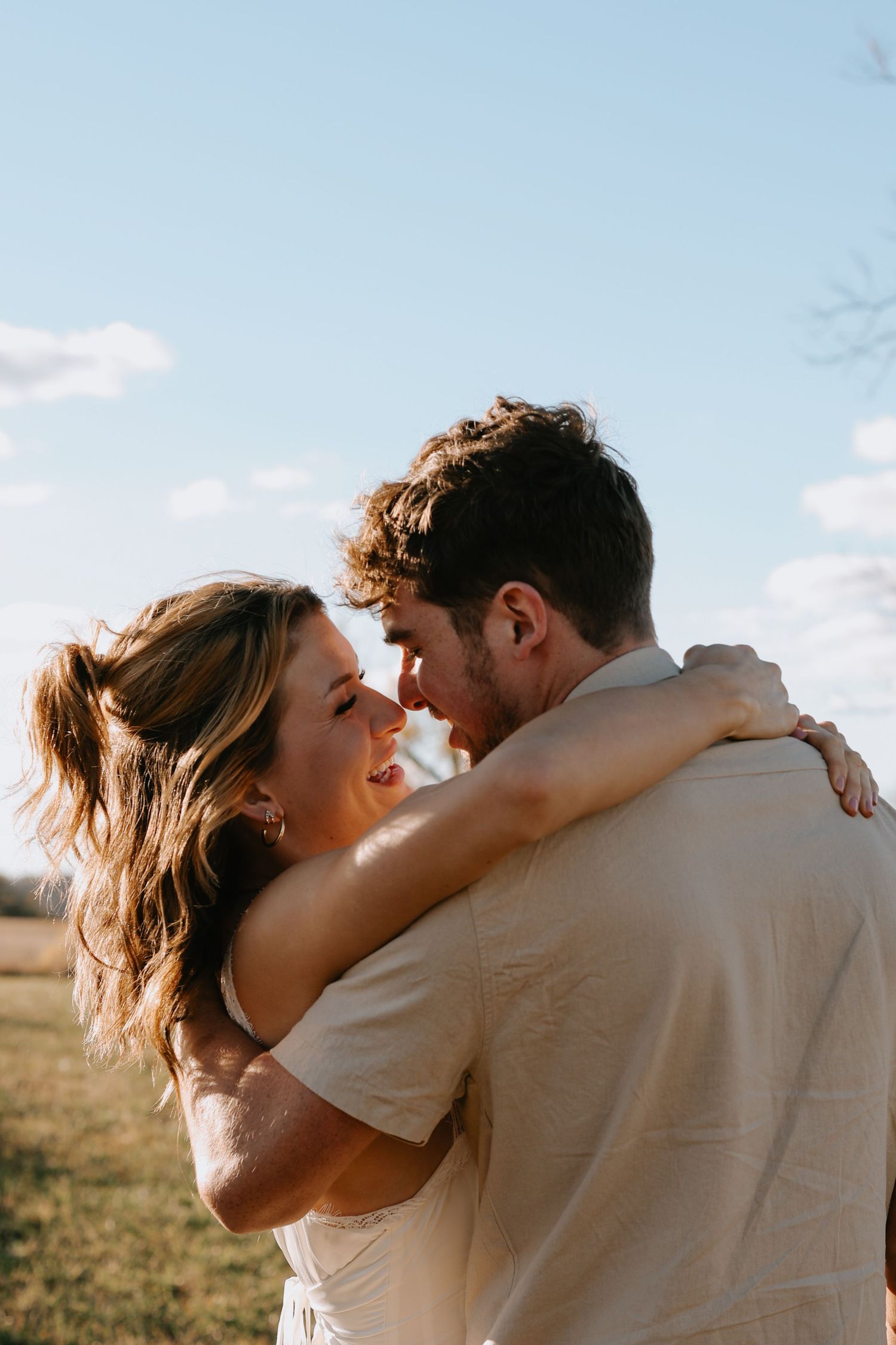 A couple embraces in a romantic moment outdoors during golden hour with a bright blue sky in the background.
