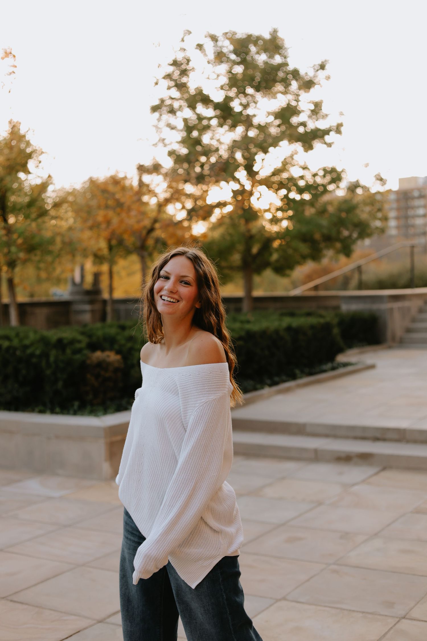 A person wearing a white off-shoulder sweater poses outdoors during golden hour in an urban setting.